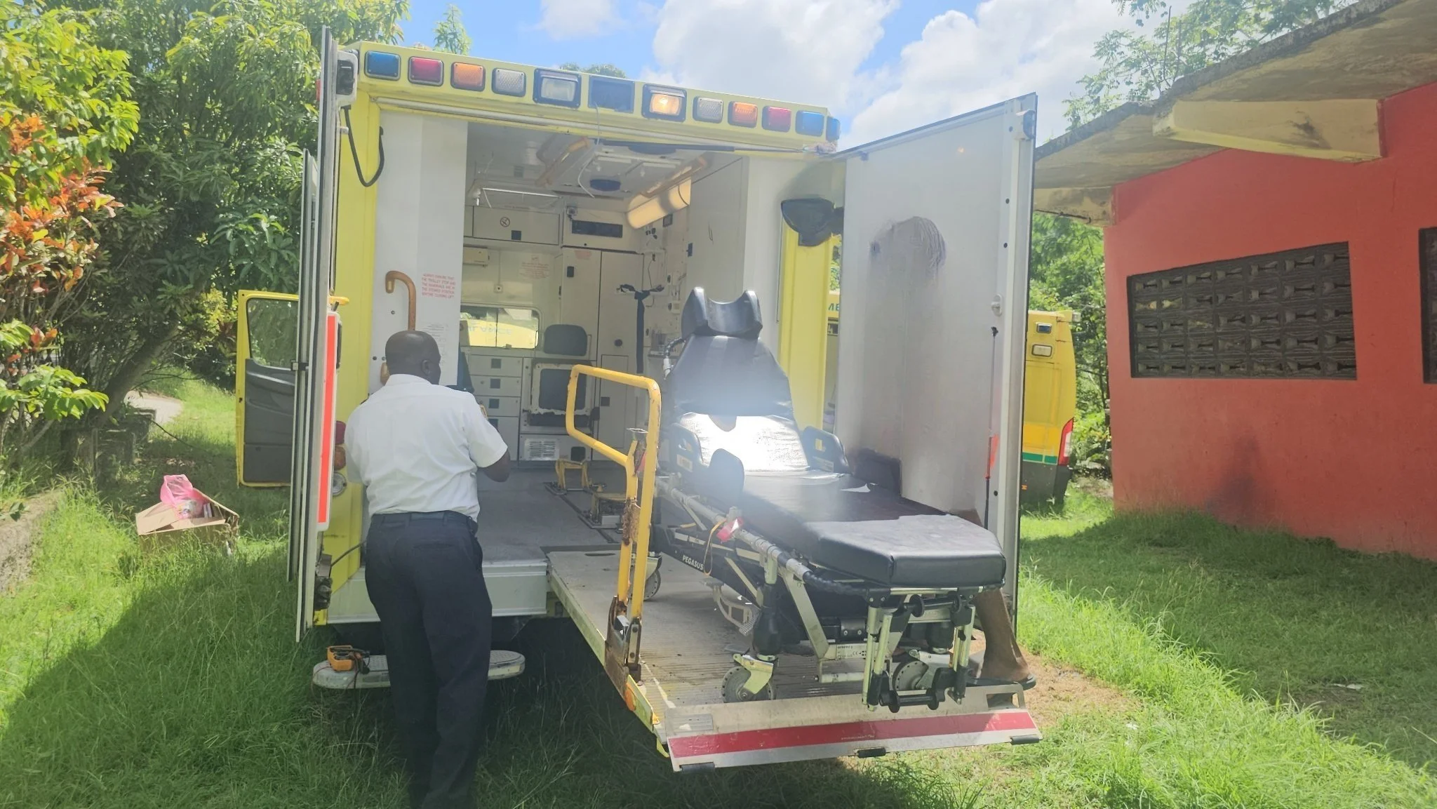A person standing outside an ambulance with its rear doors open, revealing a stretcher inside, parked on a grassy area near a red building and green trees.