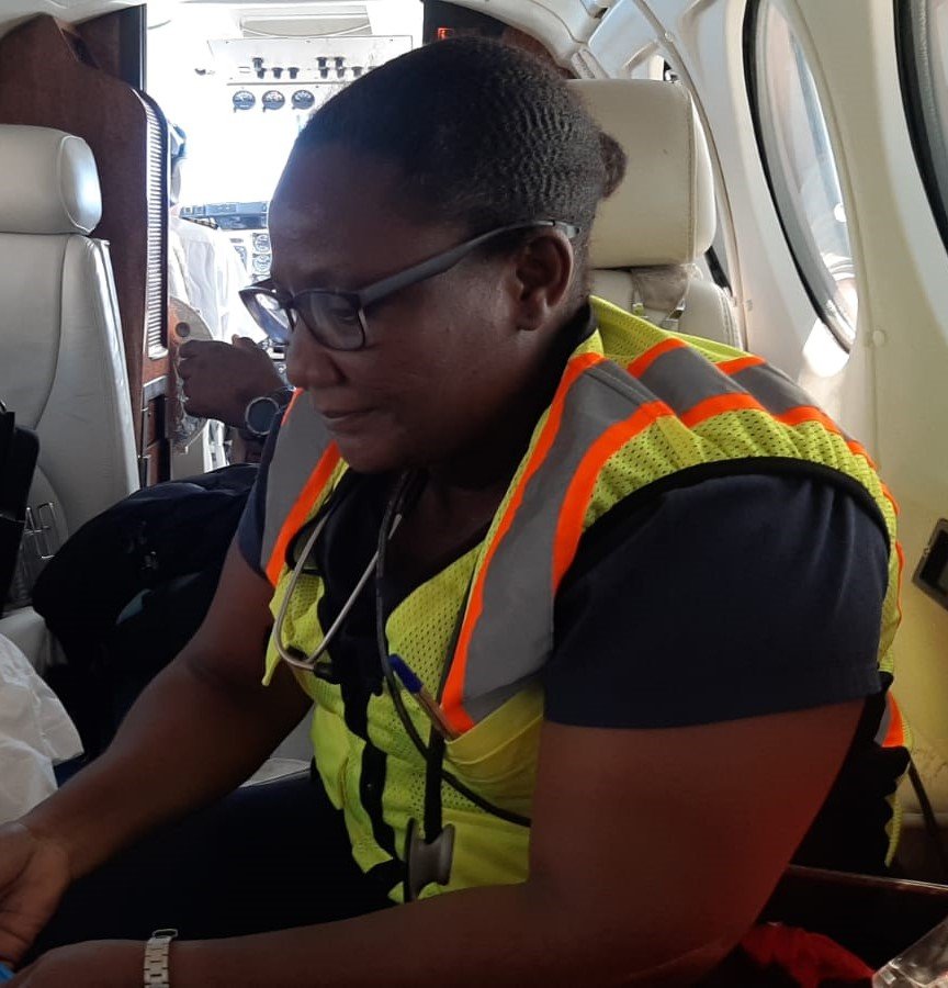 A woman wearing glasses and a high-visibility safety vest sits inside an airplane, with short hair and a focused expression. In the background, a man is seated and looking at his phone.