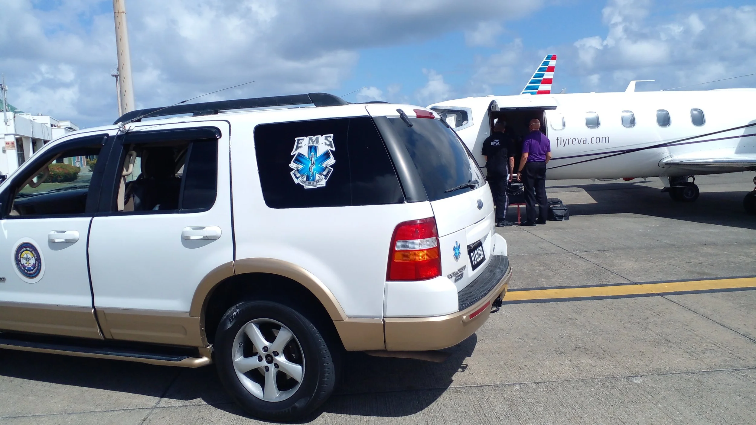An emergency medical services vehicle parked in front of a small private jet on an airport tarmac, with two people boarding the plane.