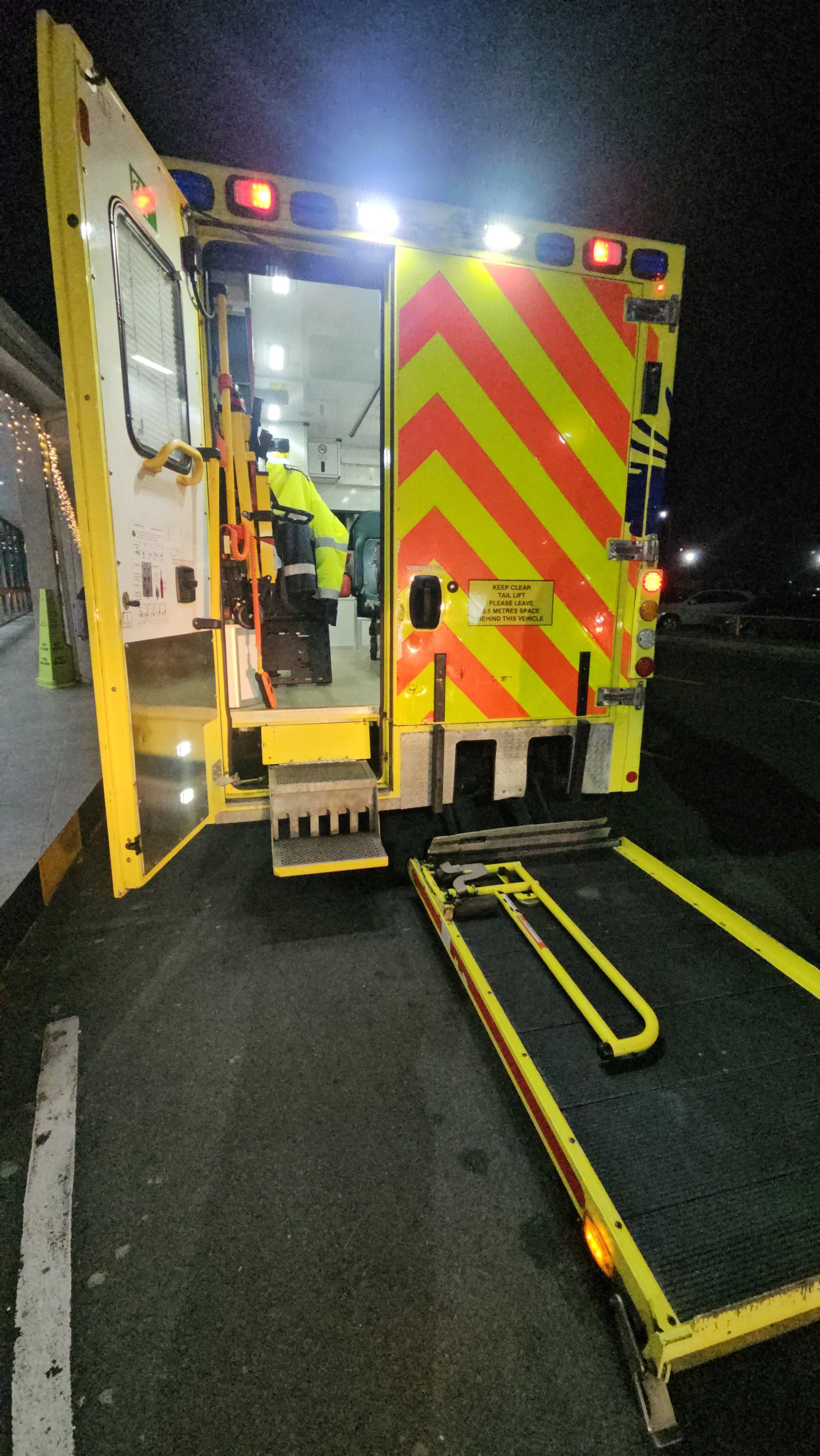 A yellow emergency vehicle with red reflective stripes and a lowered ramp, parked at night.