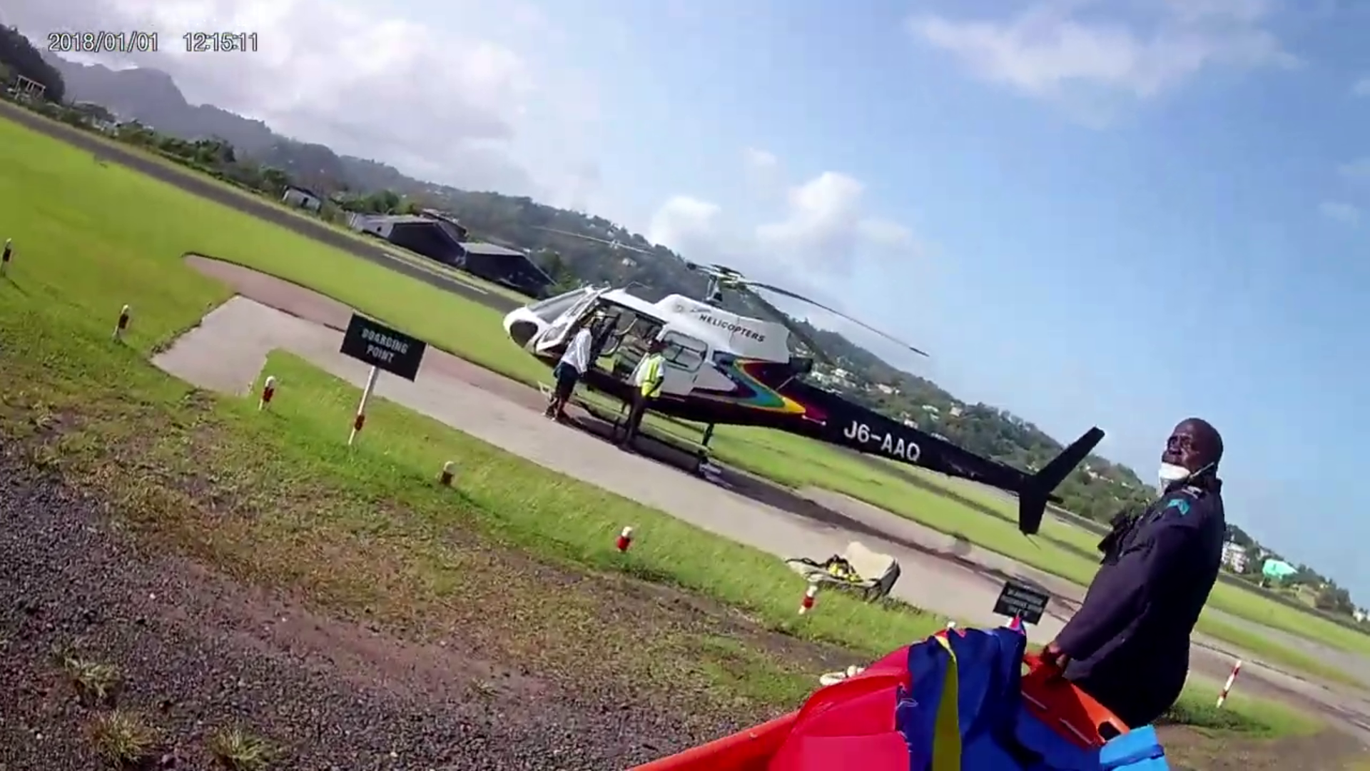 A helicopter on a tarmac with people around it, near a grassy area. A person in a uniform stands nearby with a bag, and another person enters the helicopter. The sky is partly cloudy with mountains in the background.