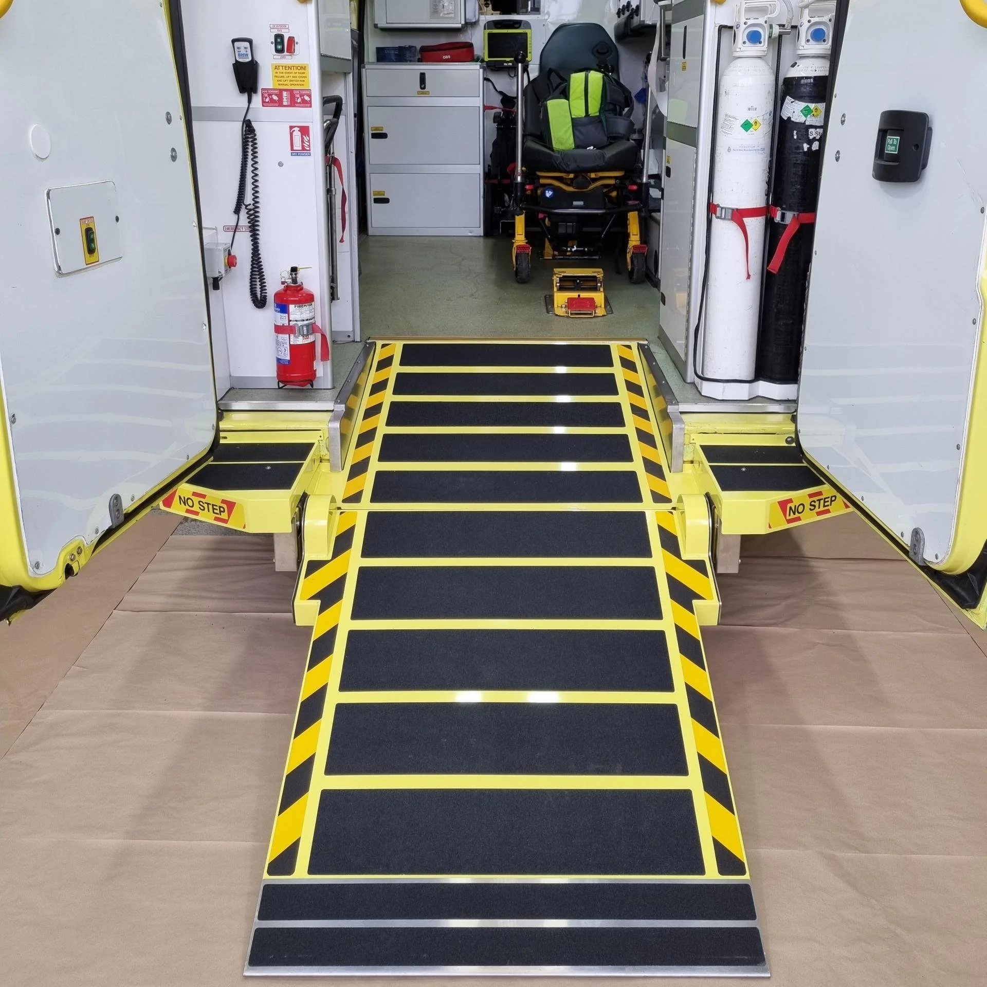 An ambulance interior entrance with a fold-out wheelchair ramp with black and yellow caution stripes, and equipment including oxygen tanks, a backpack, and medical supplies inside.
