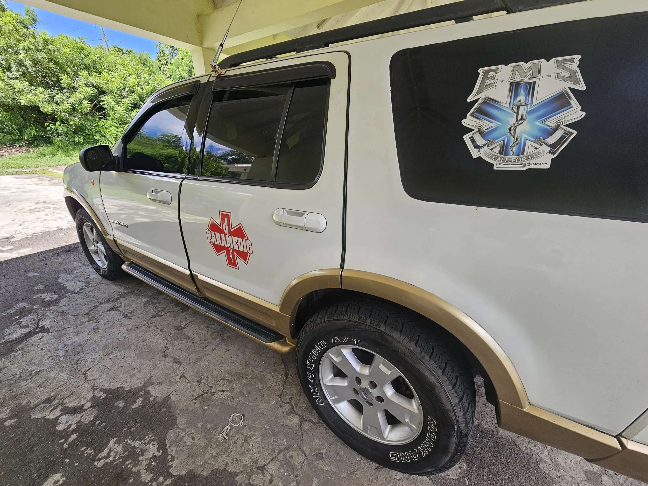 An emergency medical services (EMS) vehicle, a white SUV with gold accents, parked under a shelter. The vehicle has a red paramedic emblem on the door and a blue EMS badge with a caduceus symbol on the rear side window.