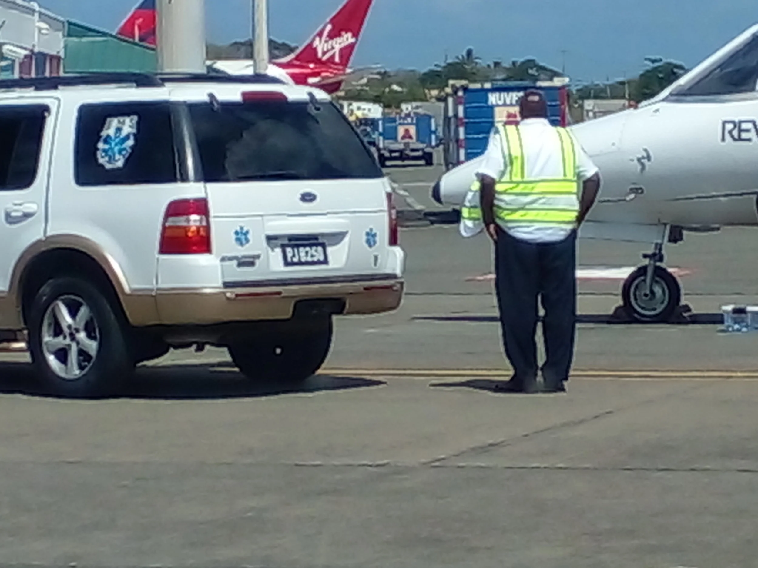 A man wearing a reflective safety vest standing near a small jet aircraft and a vehicle at an airport tarmac. Several commercial airplanes are visible in the background.