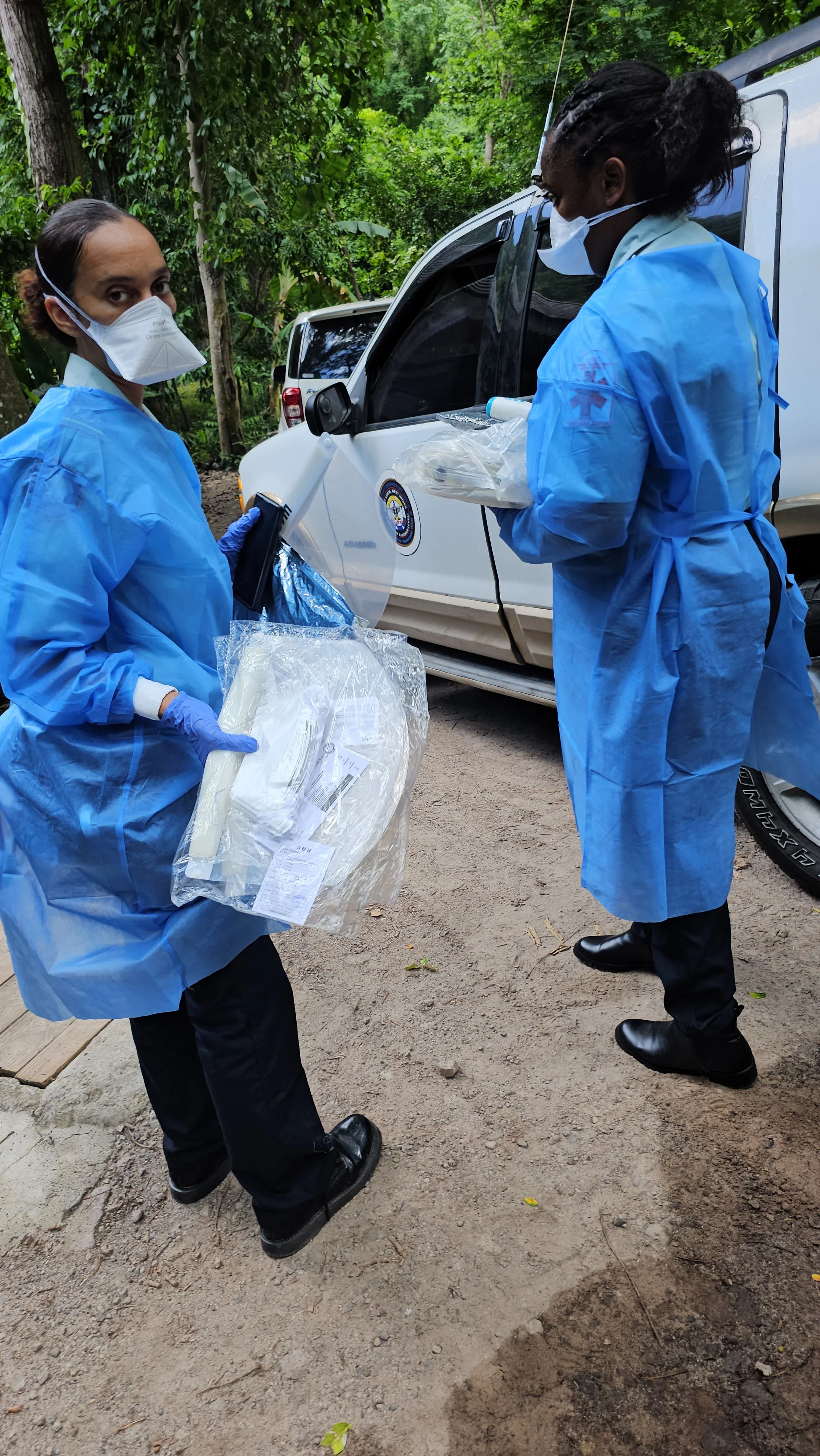 Two healthcare workers in blue protective gowns, masks, and gloves standing outdoors next to a white vehicle in a wooded area, handling medical supplies.