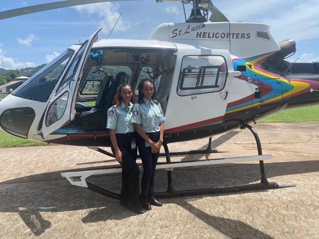 Two female flight attendants in uniform standing in front of a helicopter labeled "St. Lucia Helicopters." The helicopter is parked on a concrete surface with green grass and blue sky in the background.