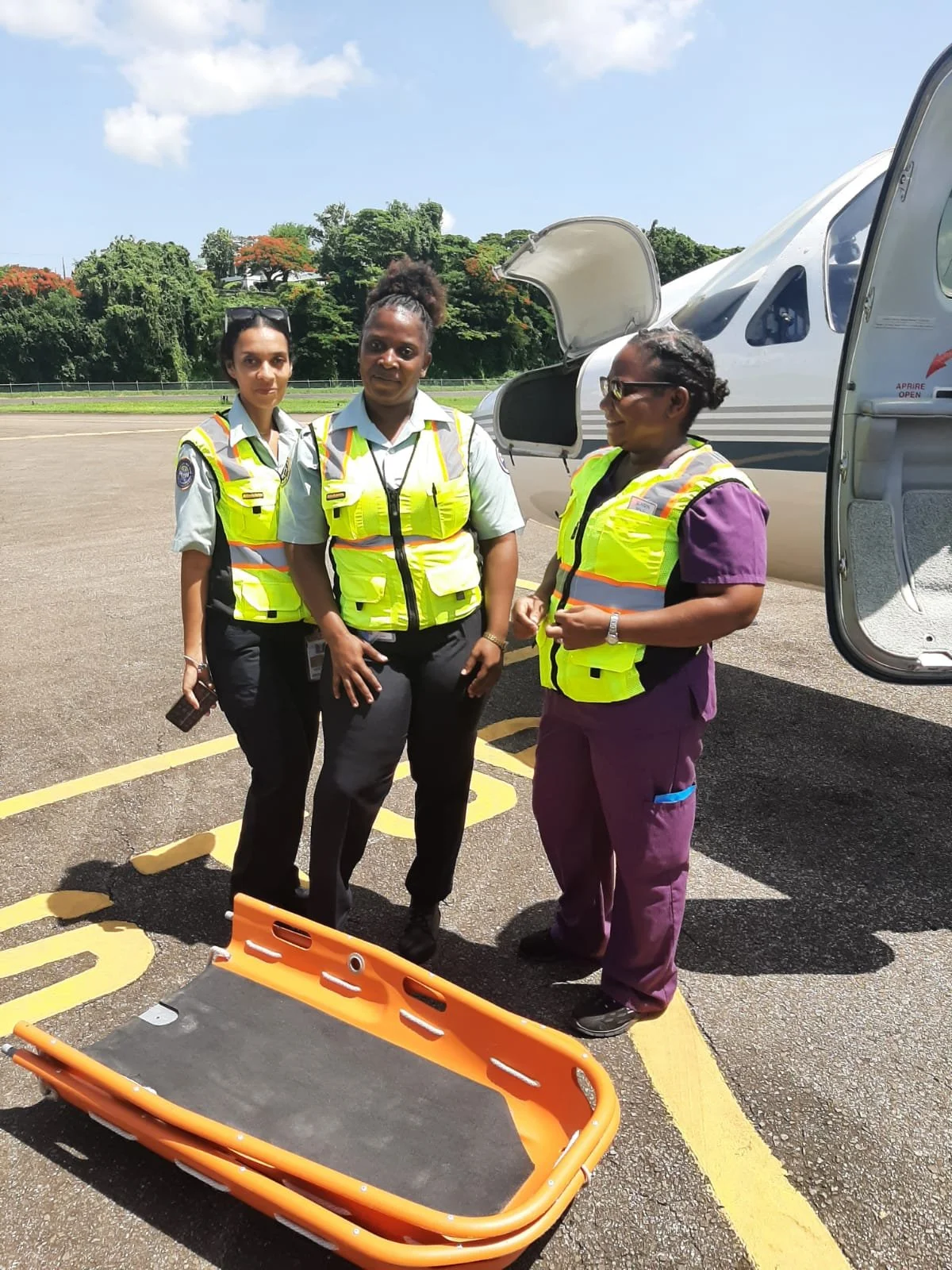 Three women standing on an airport tarmac near a small aircraft and an orange stretcher, wearing high-visibility safety vests and professional attire.