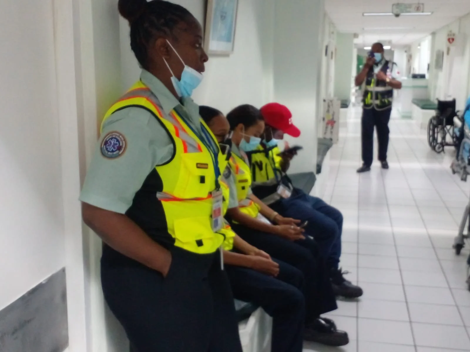 Four healthcare workers seated in a hospital corridor, wearing masks and high-visibility vests, with one standing and looking to the side. A person in the background is taking a photo, and a wheelchair is visible to the right.
