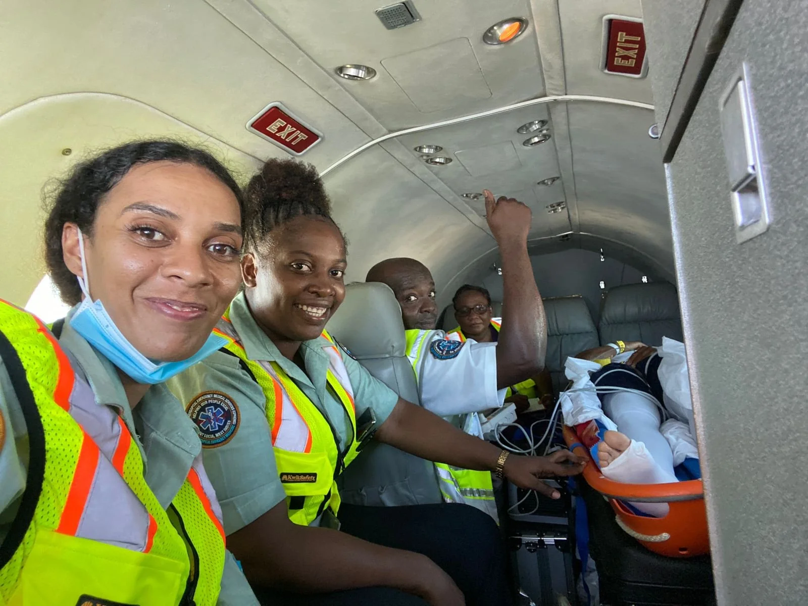 Four emergency medical personnel sitting in the aisle of an aircraft, smiling at the camera with one of them pointing to the ceiling, in front of a patient on a stretcher.