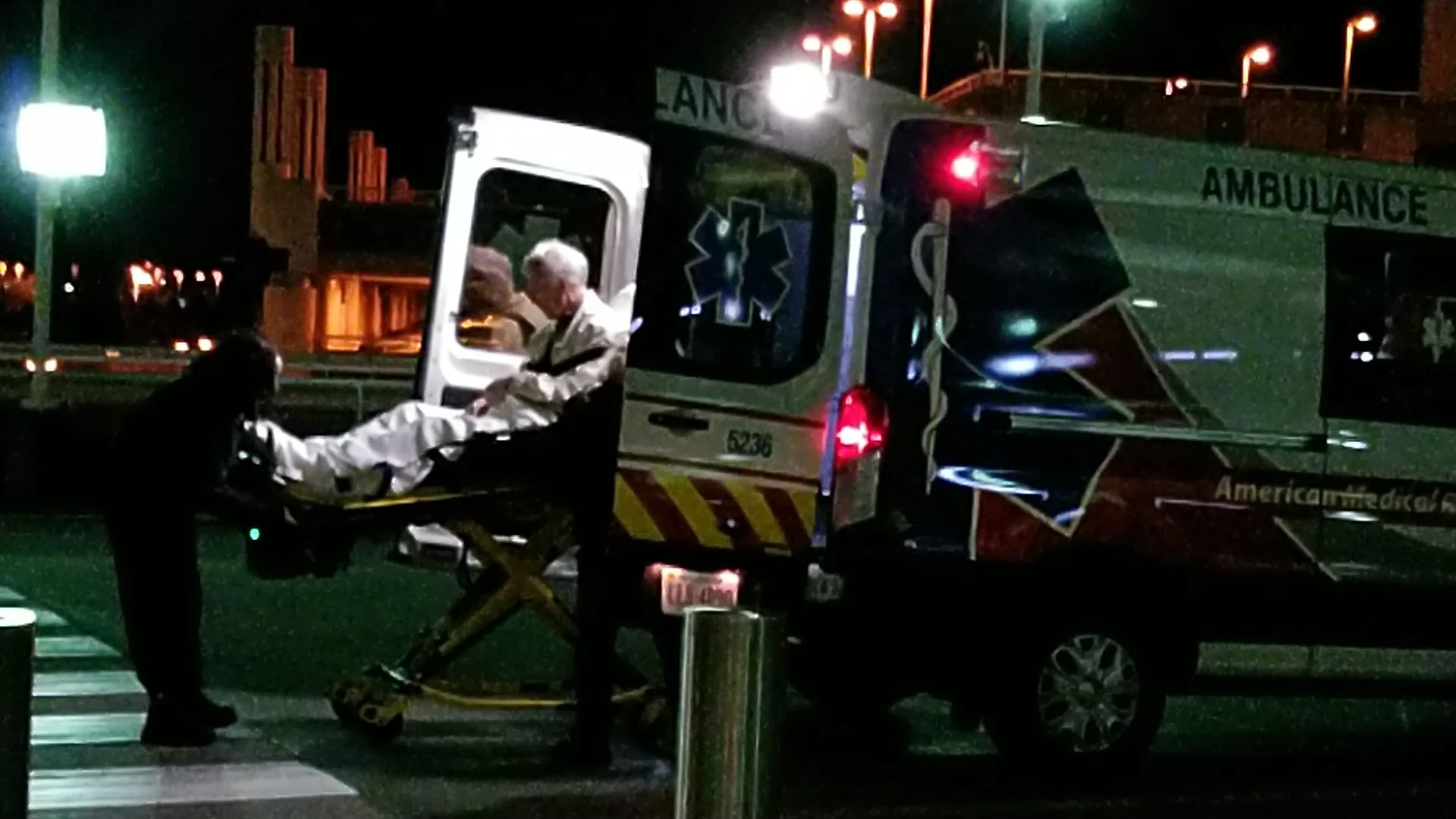 Night scene of an ambulance transporting a patient on a stretcher, with two medical personnel attending to the patient. The ambulance has emergency markings and symbols, and it is parked near a crosswalk with streetlights and urban buildings in the b