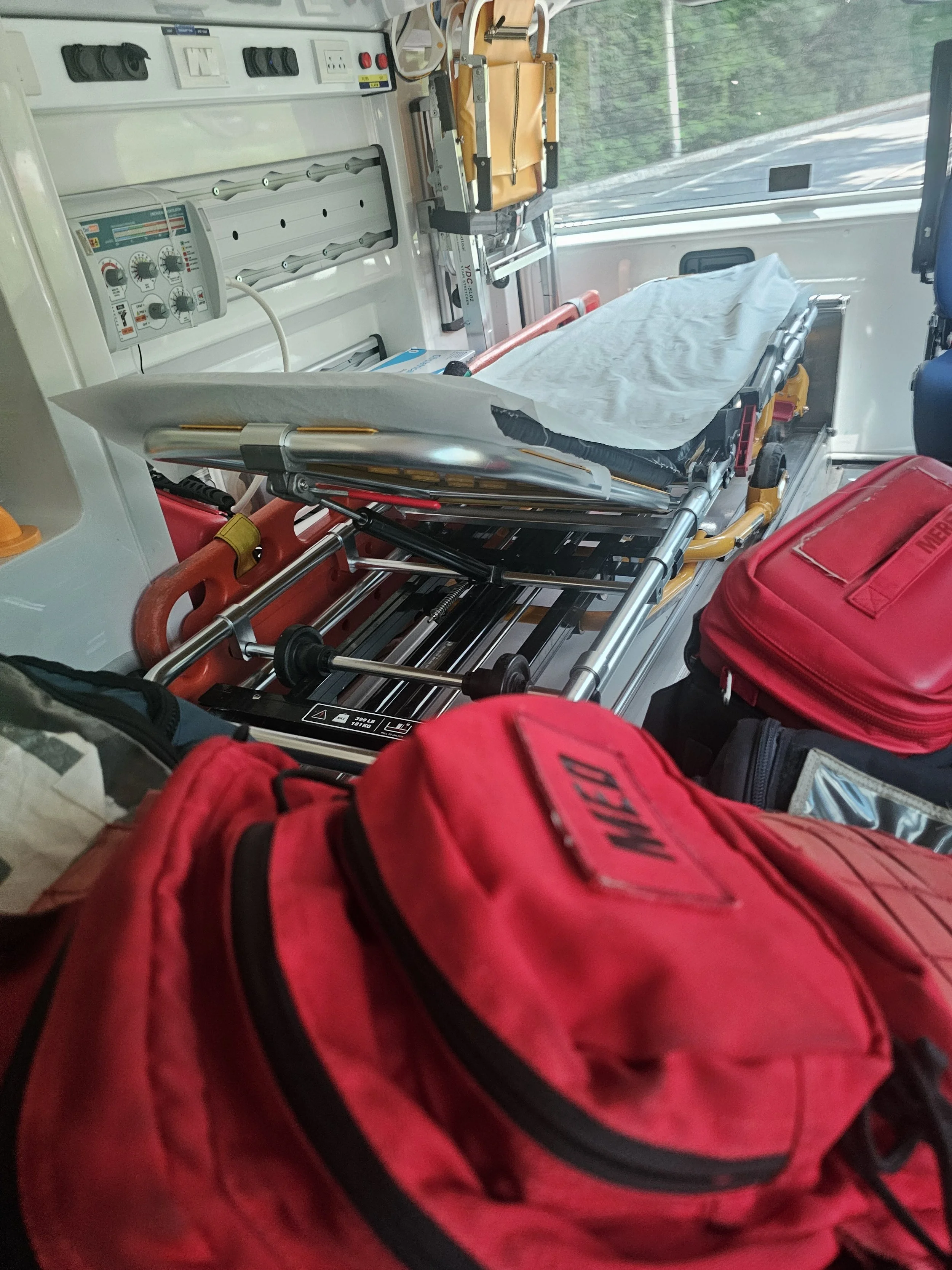 Inside an ambulance showing a stretcher with a white sheet, medical equipment, and red emergency bags.