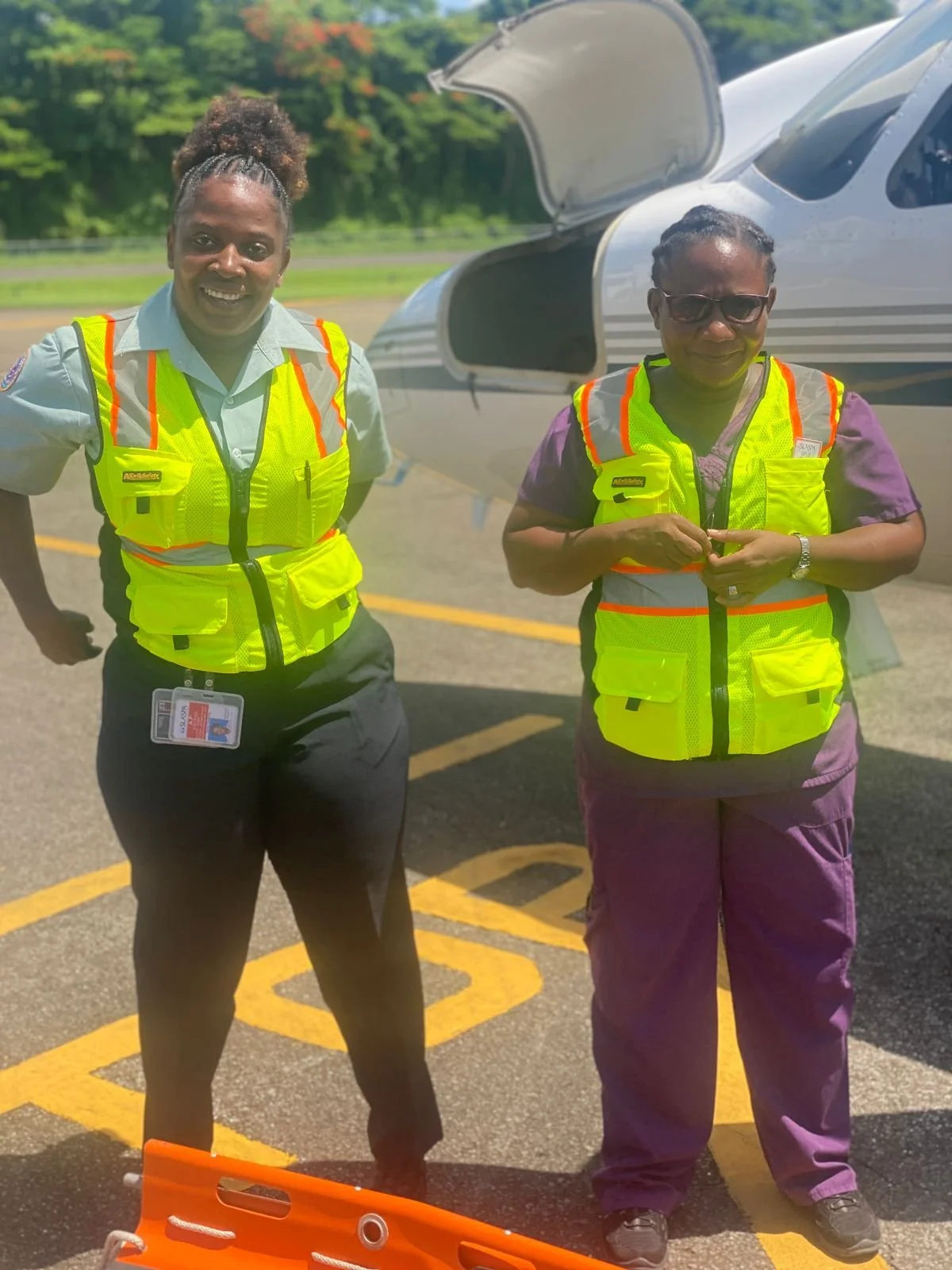 Two women standing in front of a small airplane with open cockpit, both wearing fluorescent safety vests, on a sunny day with trees in the background.