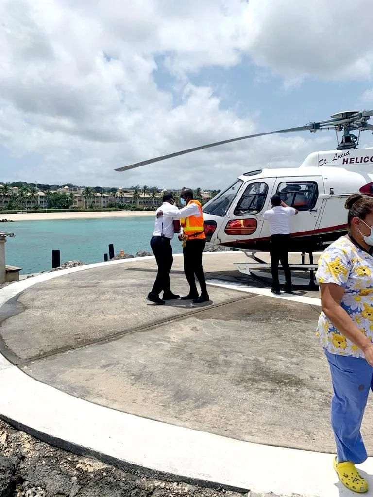 People standing near a helicopter on a helipad by the water, with one person in a reflective vest and a face mask.