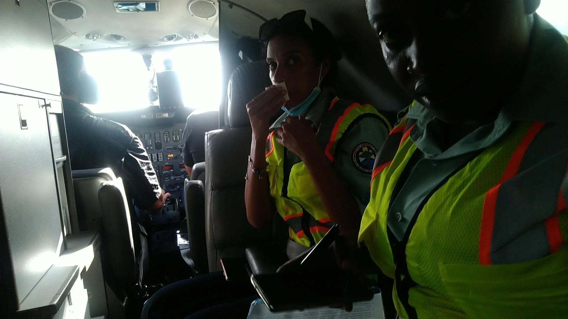 Two flight crew members, a woman and a man, inside an airplane cockpit. The woman is holding a tissue to her nose, wearing a safety vest and face mask. The man is wearing a safety vest and looking at a clipboard. The pilots are seated at the airline'
