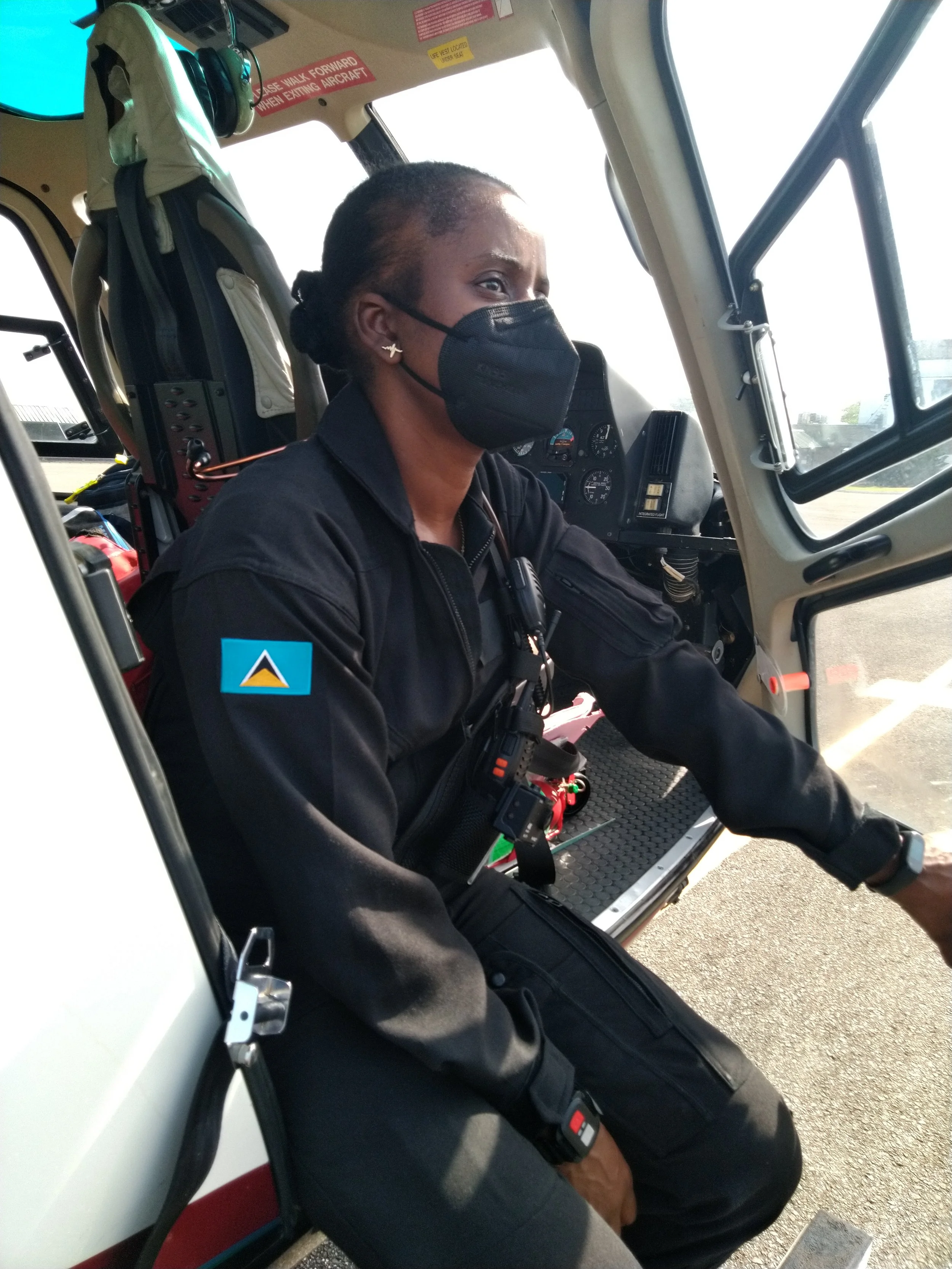 A female aviation crew member sitting in the cockpit of an aircraft, wearing a black mask, black uniform with a patch on the sleeve, and various communication equipment attached to her uniform.