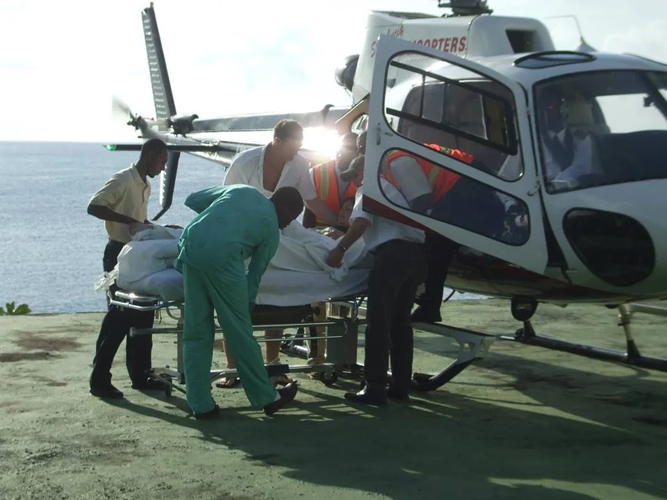 Medical personnel loading a patient into a helicopter on a shoreline or dock, with the ocean in the background.