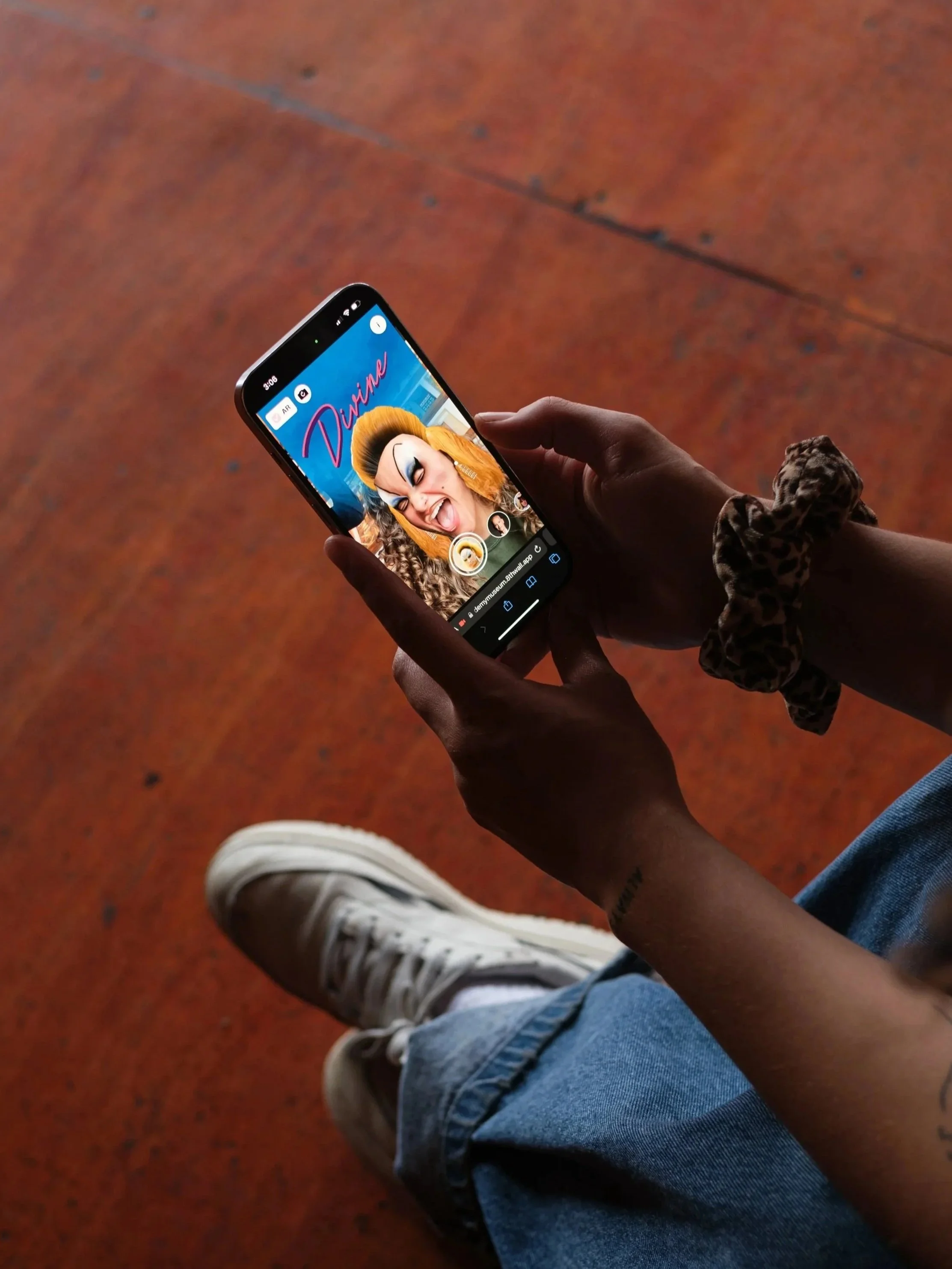 Person holding a smartphone with a photo filter of a woman with big hair and makeup, and the text 'Divine' in pink. The person's legs and shoes are visible, with a red-orange floor in the background.