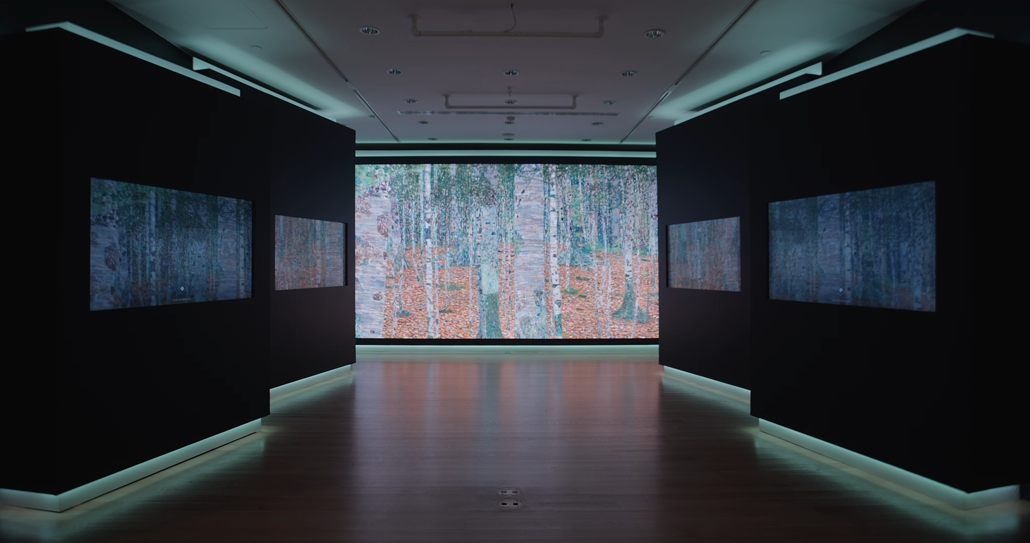 An art gallery with dark walls and wooden flooring, featuring a large digital screen displaying a forest scene with trees and falling leaves.