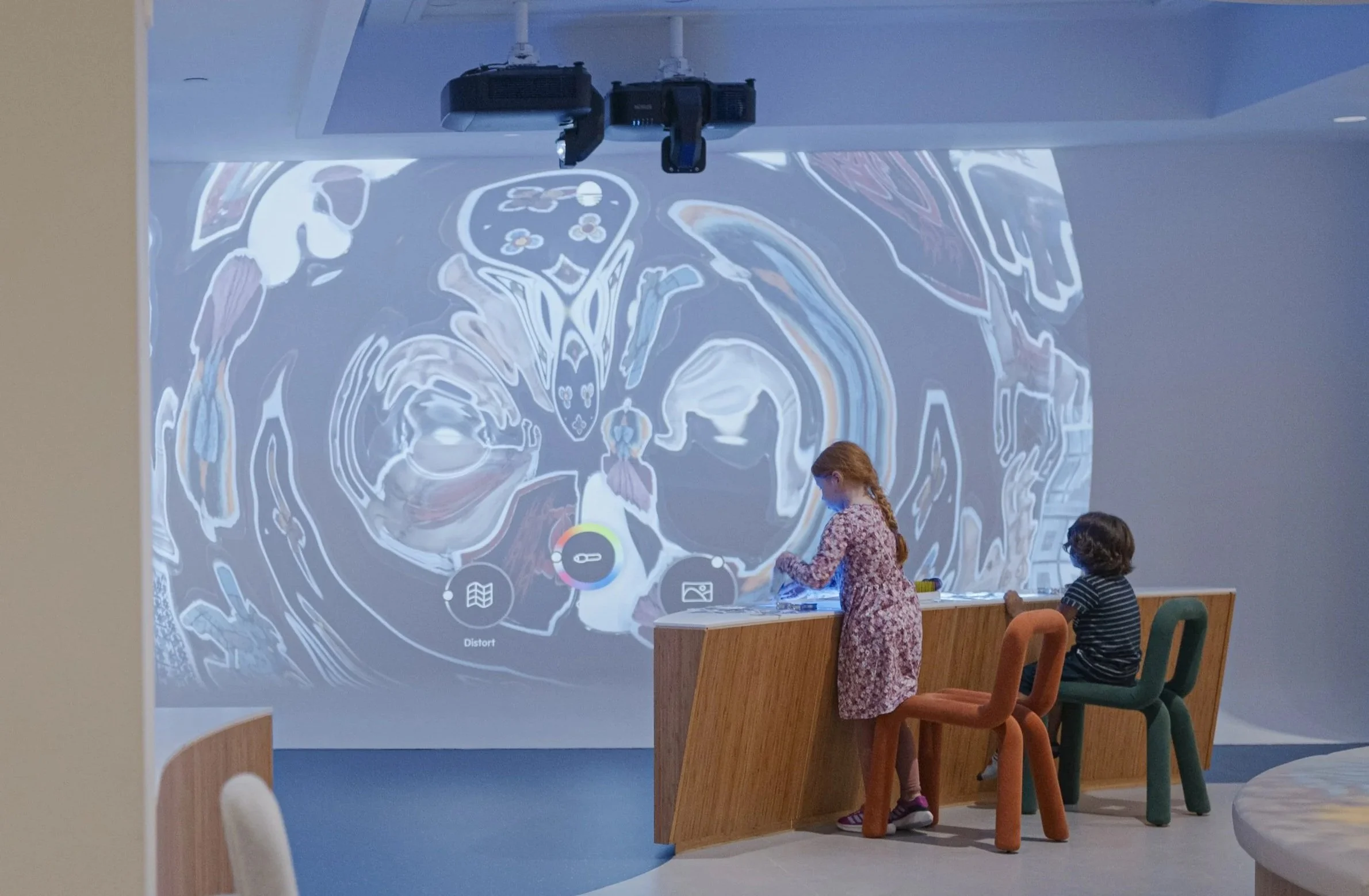 Two children, a girl in a floral dress and a boy in a striped shirt, interact with a large interactive digital display wall in a modern room. Overhead projector shows colorful abstract patterns on the wall.