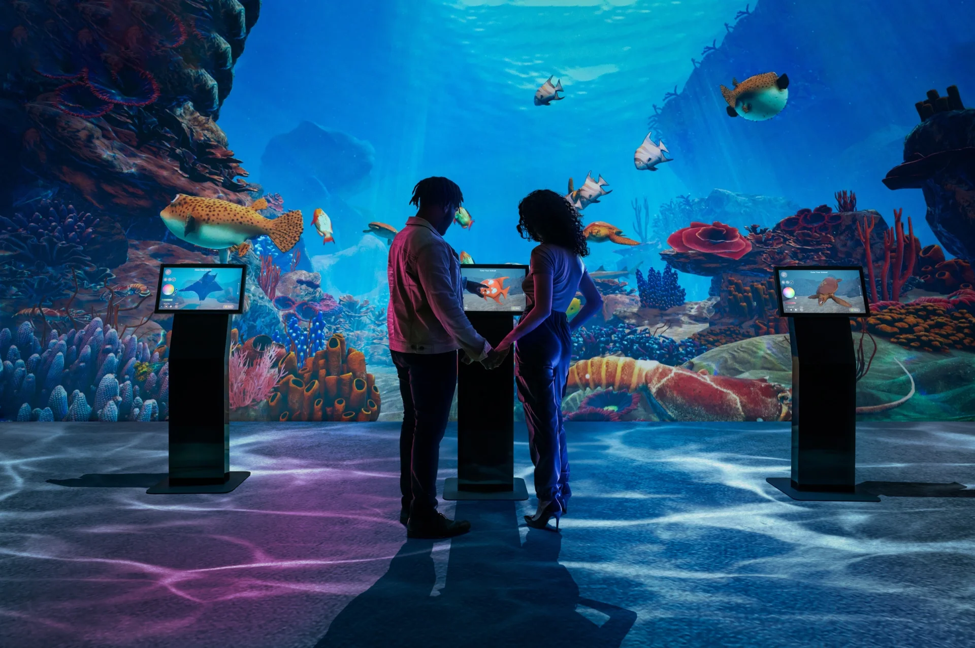 A couple stands holding hands in front of a large underwater aquarium exhibit, surrounded by colorful fish, coral, and sea creatures, with interactive screens on stands nearby.