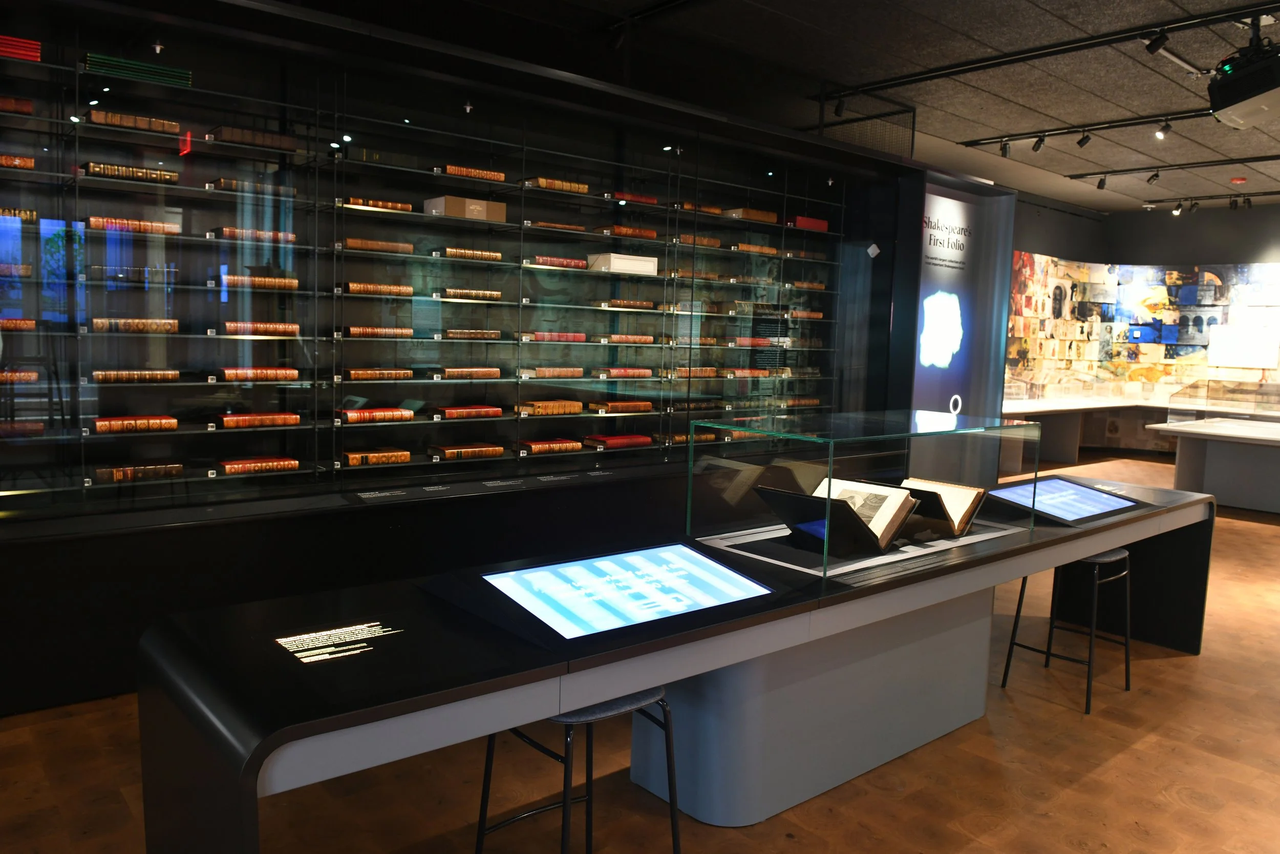A library exhibit with glass display case filled with books and digital screens on a black table in a museum setting.