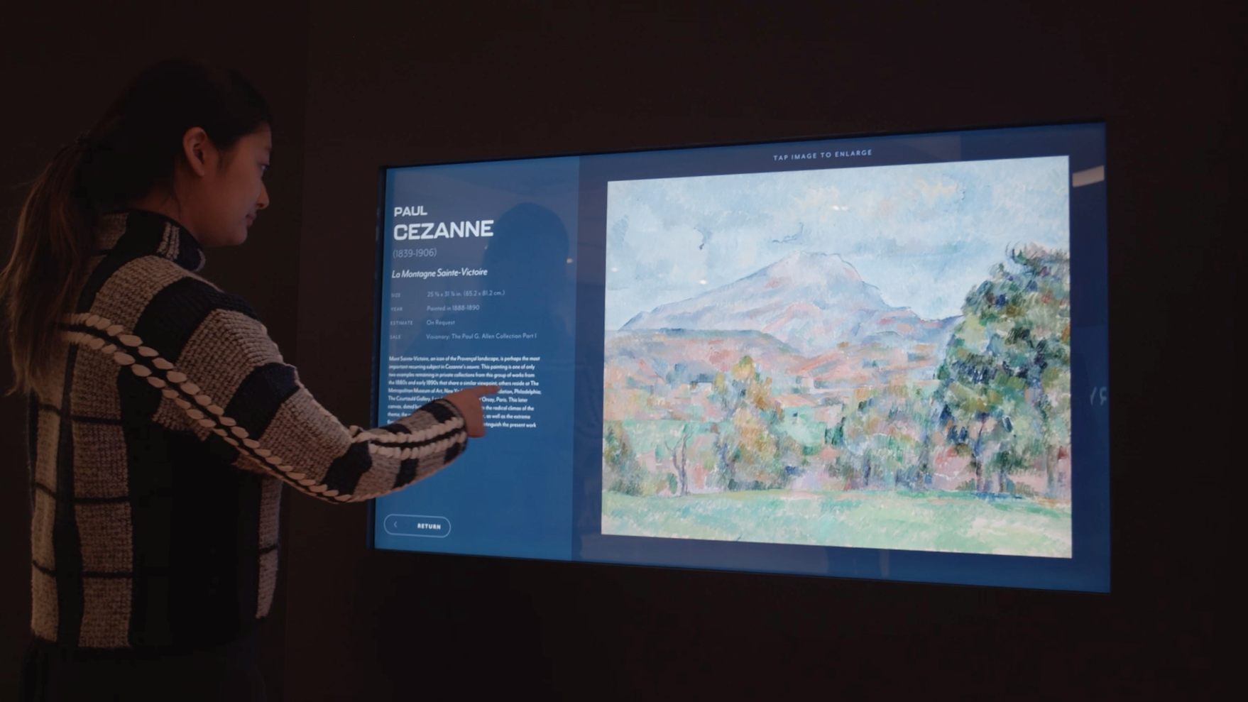 A woman in a striped sweater interacts with a large digital display showing information about the painting La Montagne Sainte-Victoire by Paul Cezanne.