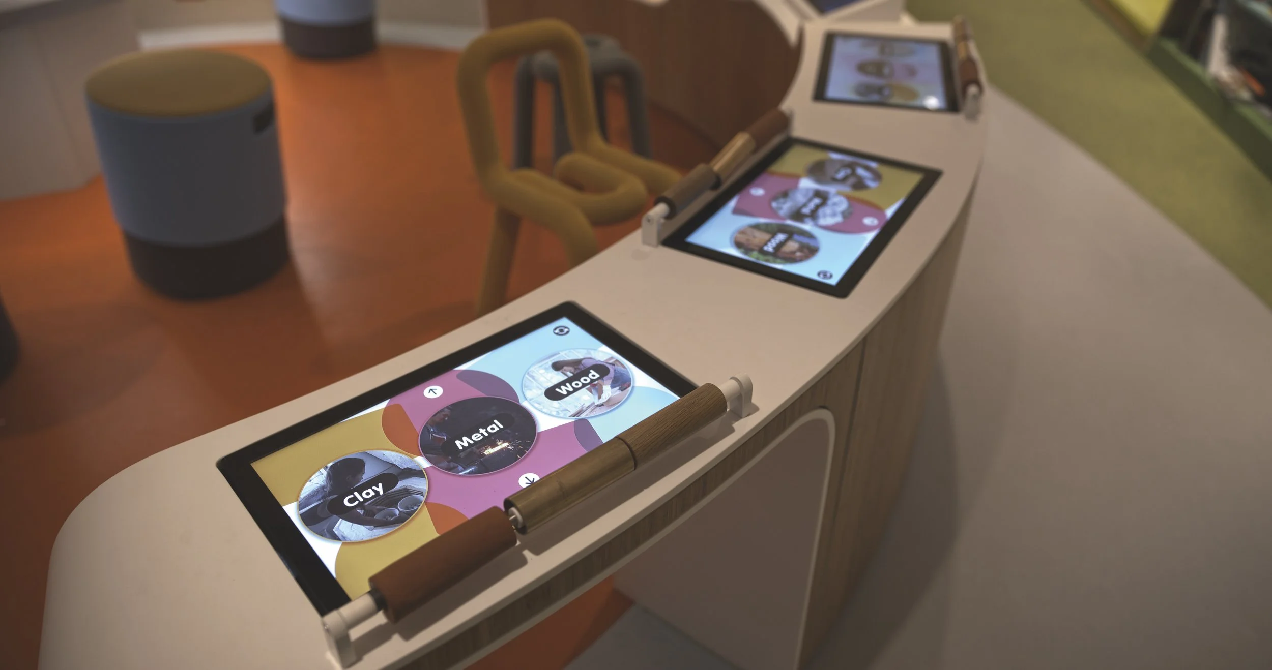 Interactive display screens on a curved white countertop with wooden handles, showing options for material types like clay, metal, and wood in a modern showroom or store. A wooden chair and a cylindrical object are visible in the background.