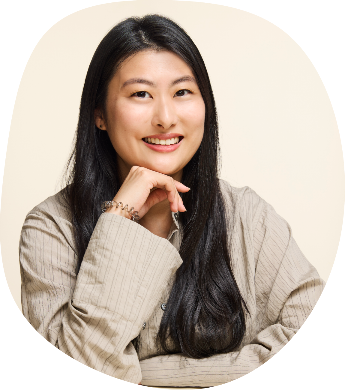 A young woman with long black hair smiling, resting her chin on her hand, wearing a beige striped shirt and a silver bracelet, against a light background.