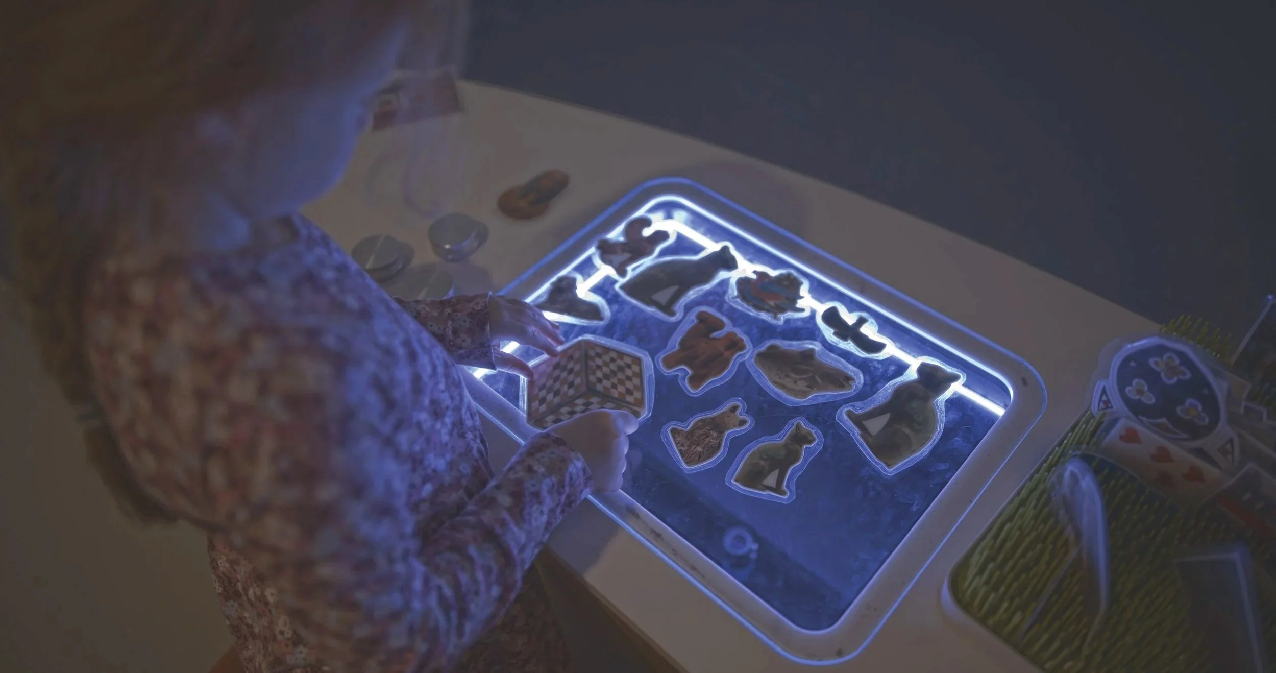 Child playing a digital game with cutout puzzle pieces on a backlit touchscreen table, surrounded by digital game accessories.