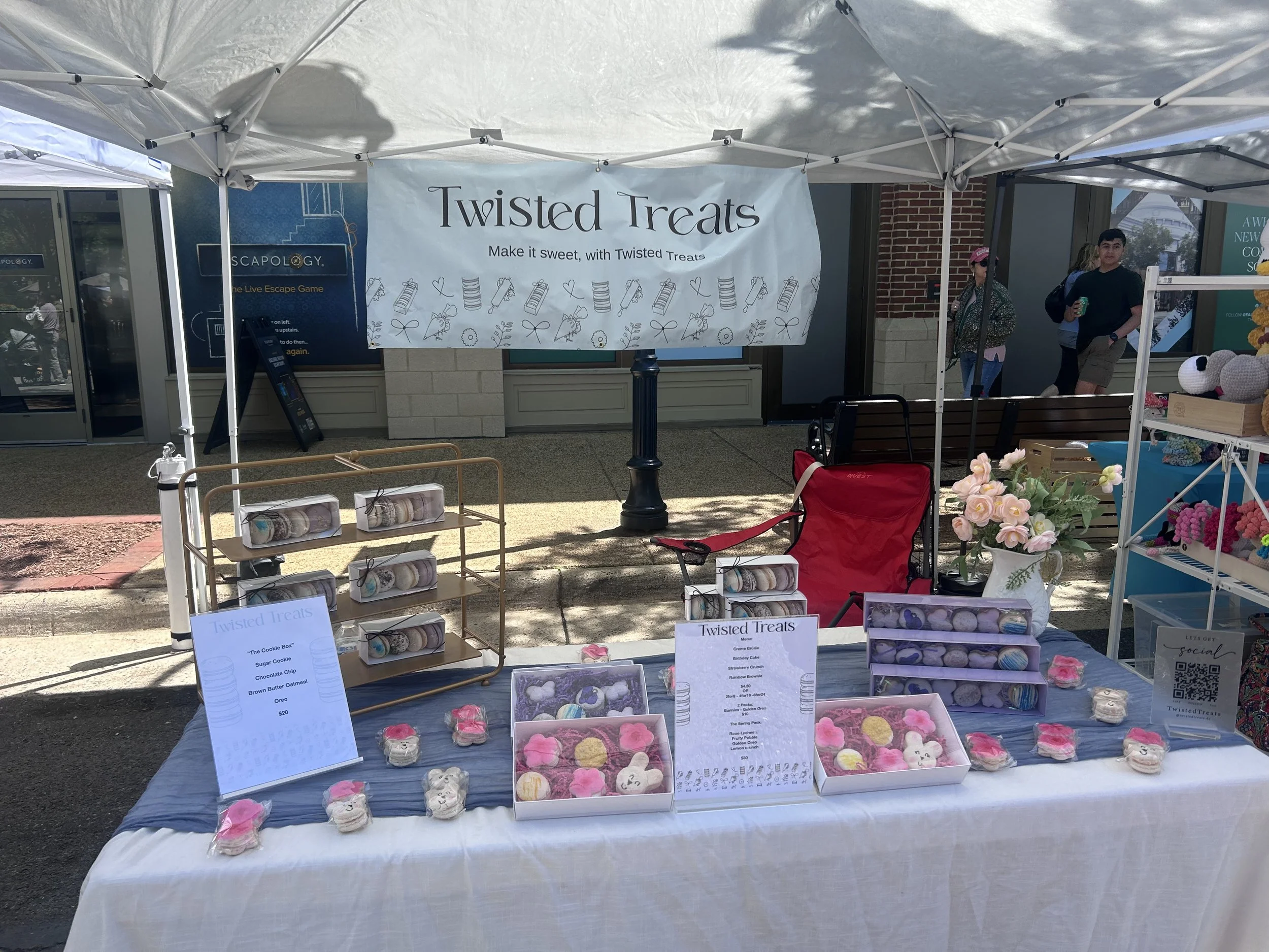Outdoor market stall with a sign that reads "Twisted Treats" selling decorated cookies in various shapes and colors. Background shows a few people strolling, a brick building, and other market booths.