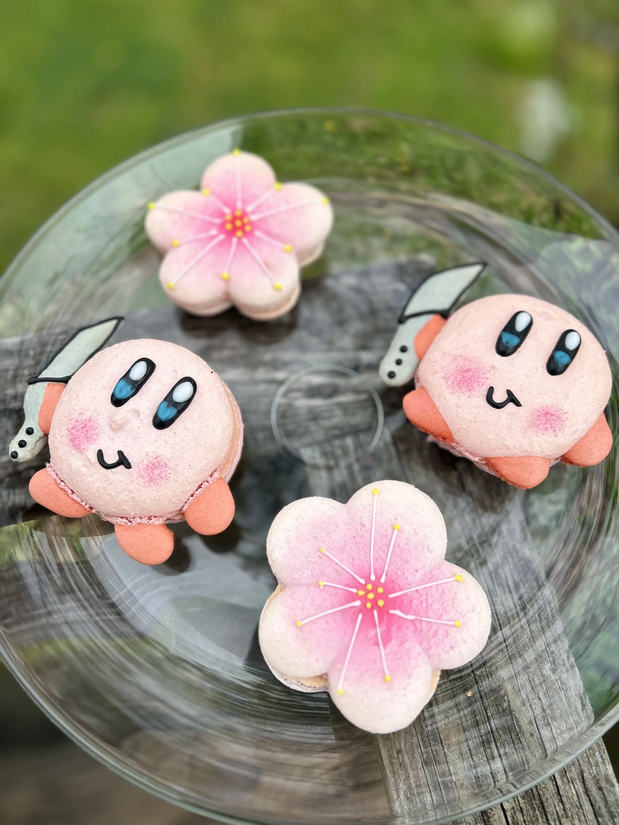 Two adorable pink bunny-shaped macarons with blue eyes and blushing cheeks, placed beside two pink flower-shaped macarons with yellow stamens on a glass plate, outdoors on a wooden surface.