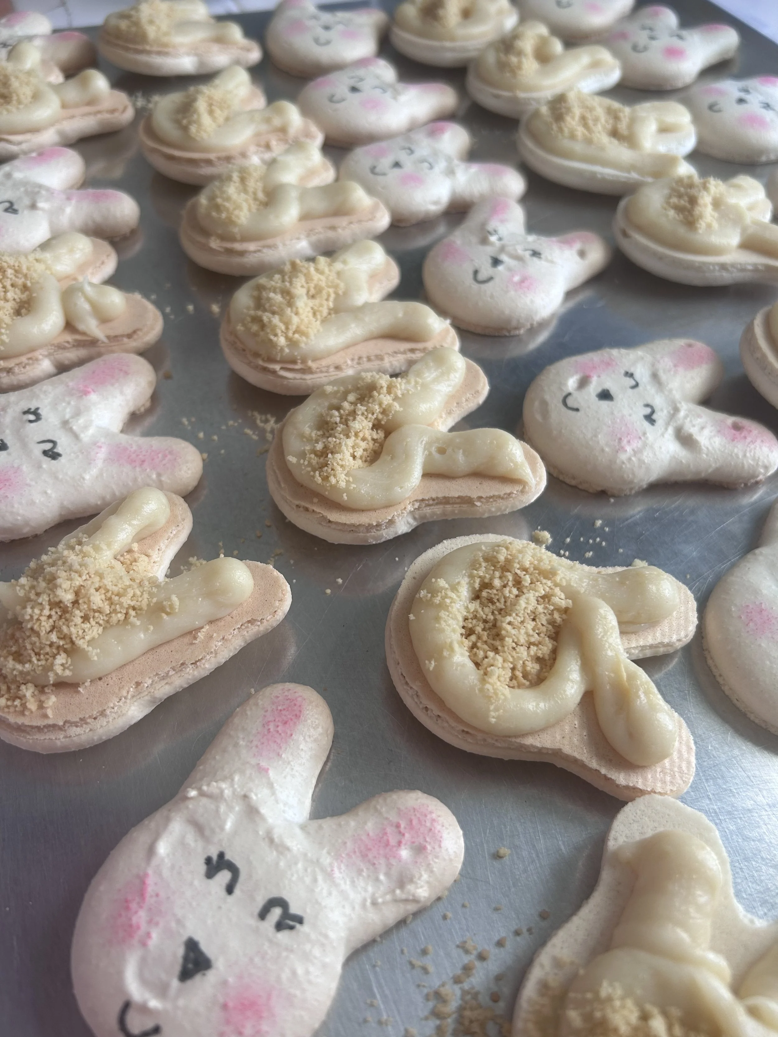 Sugar cookies shaped like bunnies with white icing, pink cheeks, black eyes, and nose, decorated with white chocolate and crumbled cookies.