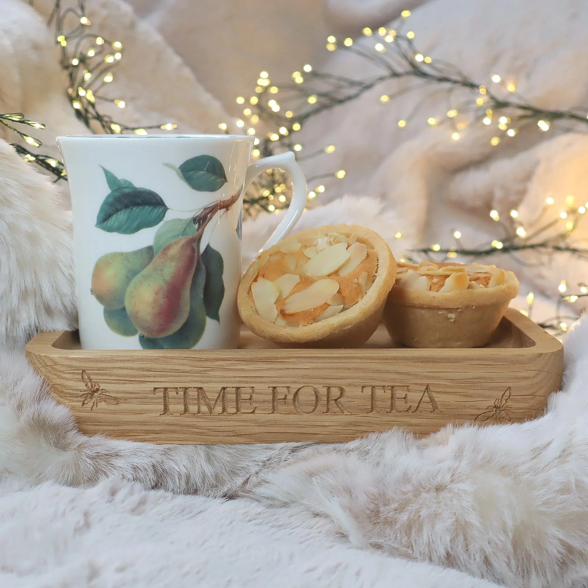 A side view of an engraved oak teatime tray with a mug of tea and mince pies housed on it with a foreground and background of faux fur in soft taupe with fairy lights.