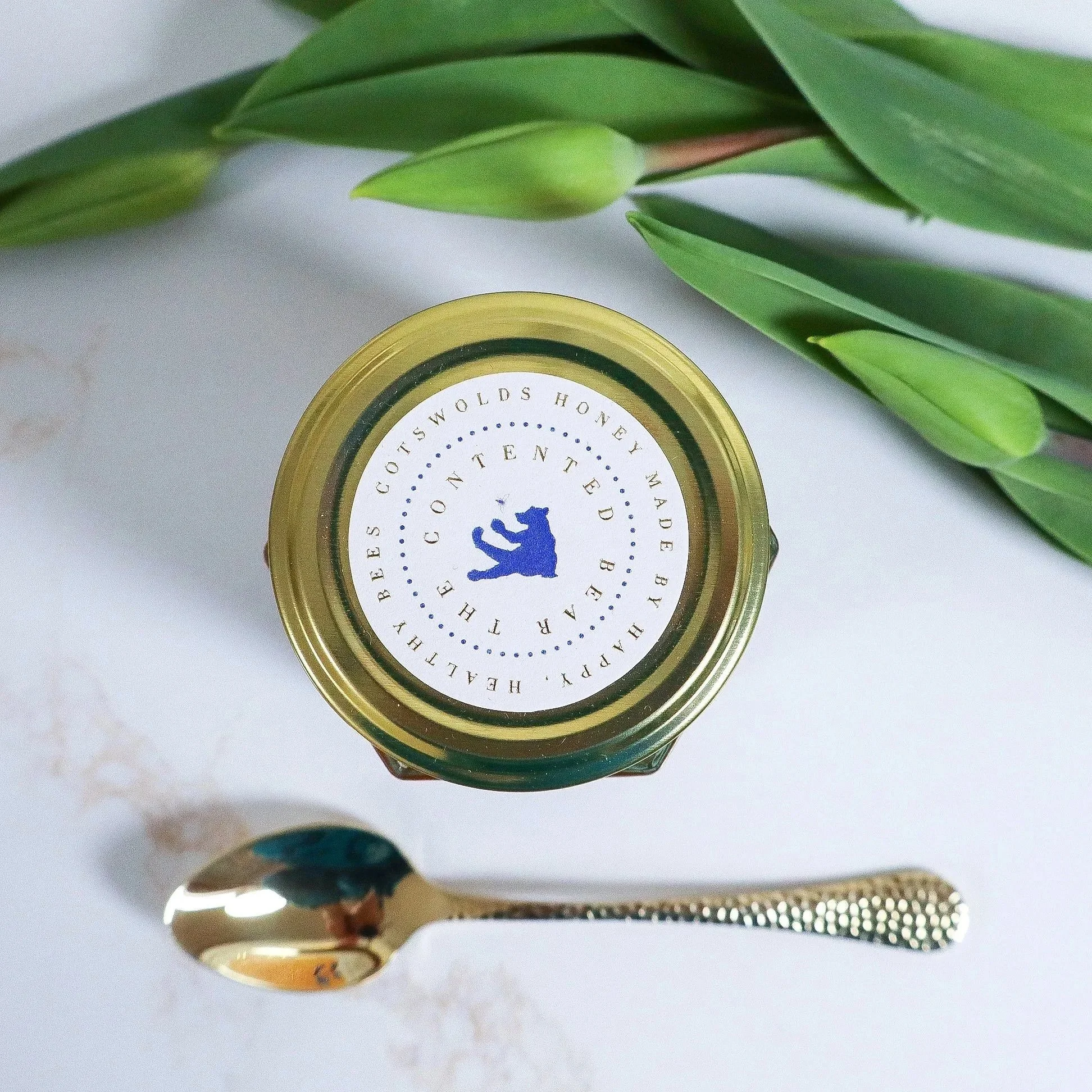 Aerial view of a luxury, sustainable jar of honey with a luxury lid label with a blue bear in profile. An arc of tulips frames the jar with a gold teaspoon sitting by the jar.