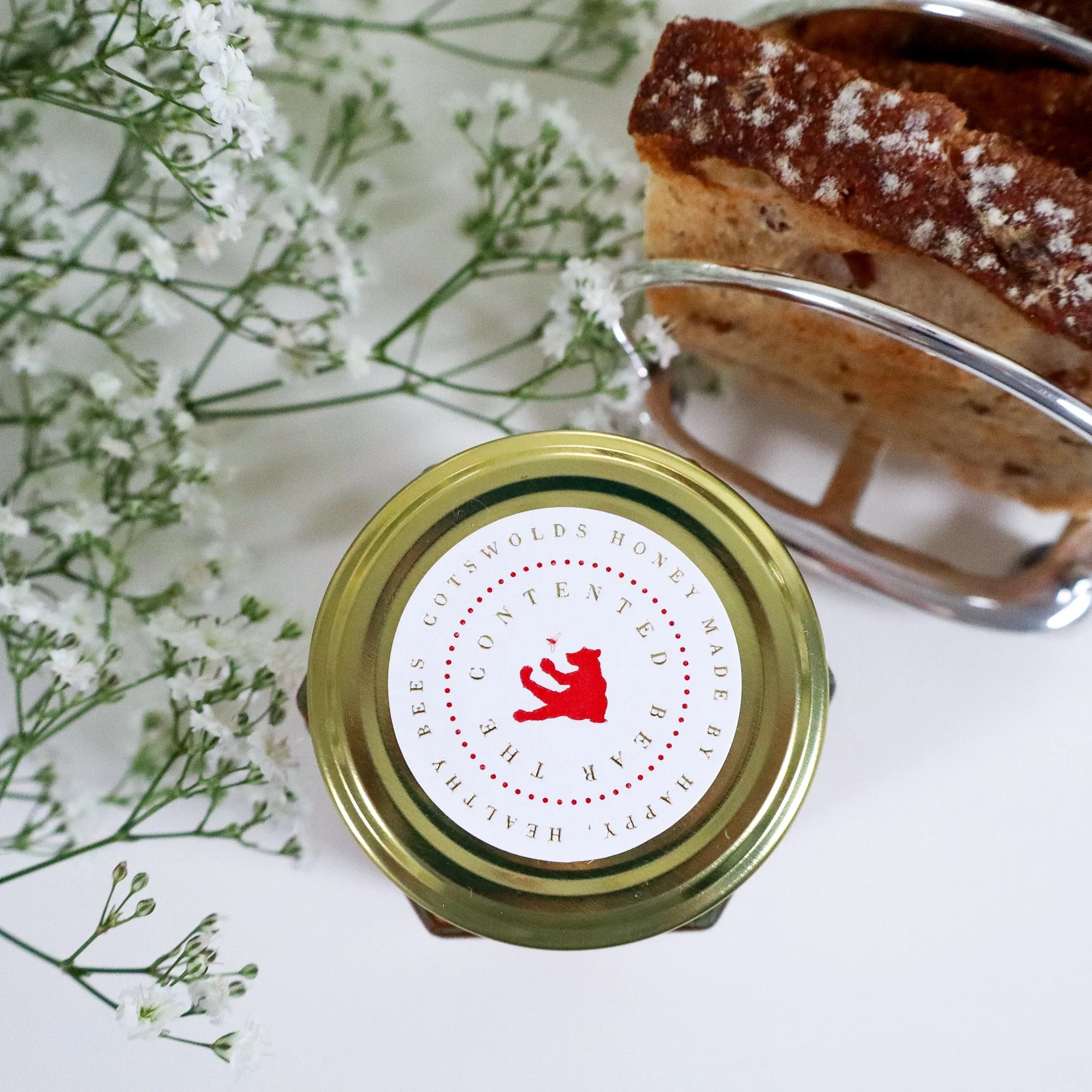 Birds eye view of the top of a luxury, ethical jar of honey with white flowers to the left and artisan toast in a silver toast rack on the right.