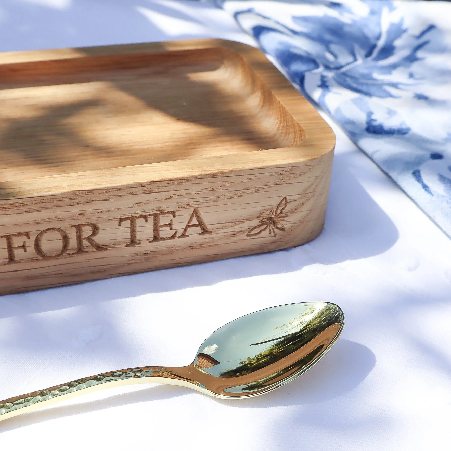 Close up of engraved solid oak teatime tray in dappled light. Alongside the tray is a golden teaspoon and bkue and white napkin.