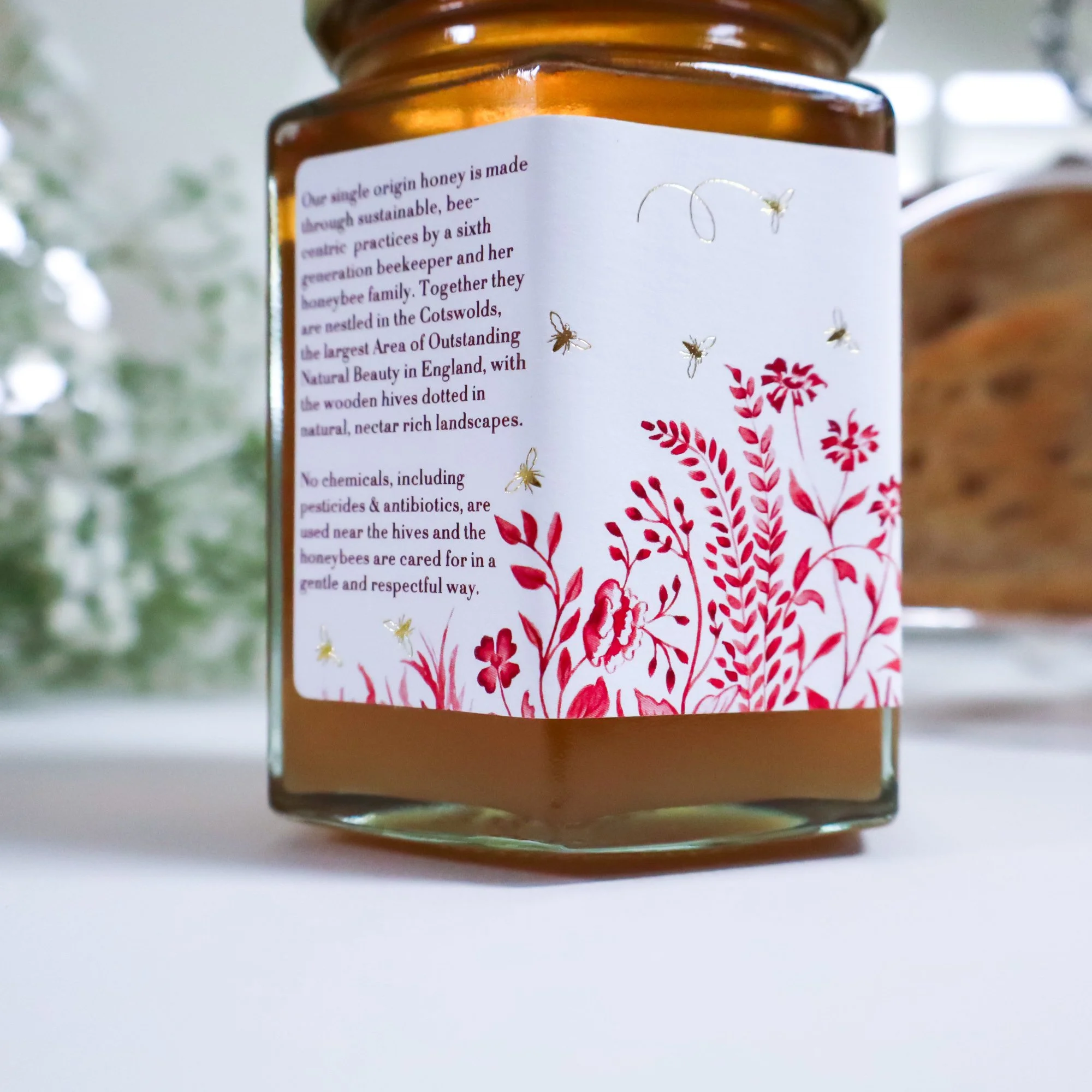 Side view of a sustainable jar of raw honey with a luxury hand illustrated label featuring a red meadow and sun with gold foil honeybees and text.