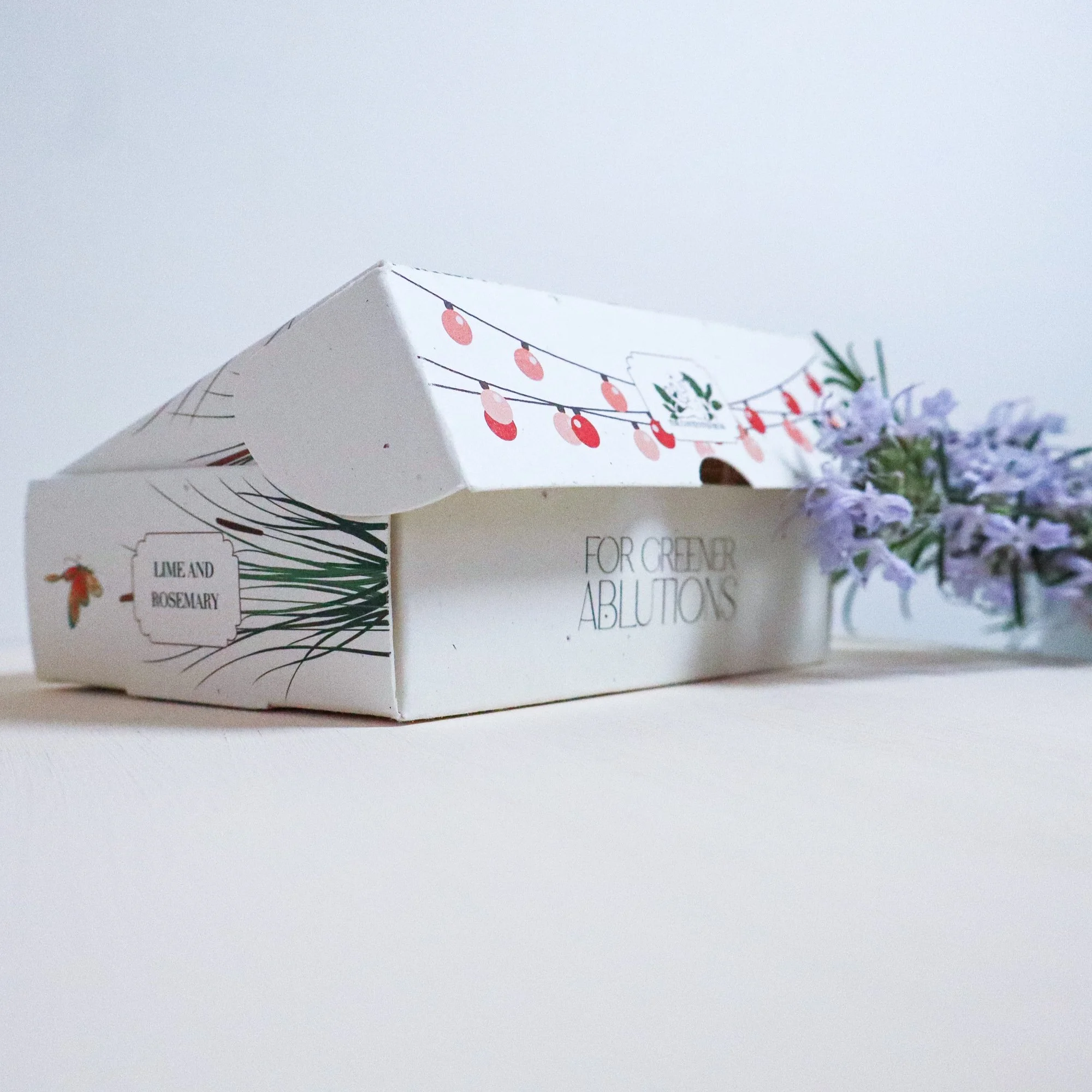 An illustrated soap box with the lid slightly open to rteveal more illustrations on. the side of the recycled card box.  A bunch of fresh flowering rosemary lays to the right of the artisan soap box.