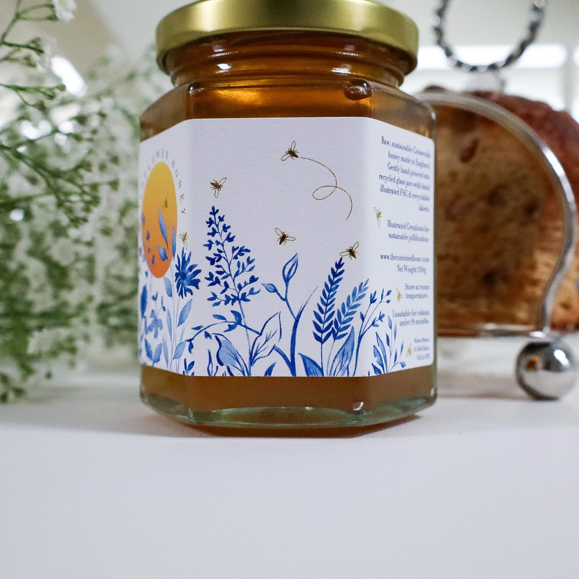 Close up side view of a beautiful, illustrated honey label on a jar of sustainable honey. A silver toast rack and white flowers are in the background.