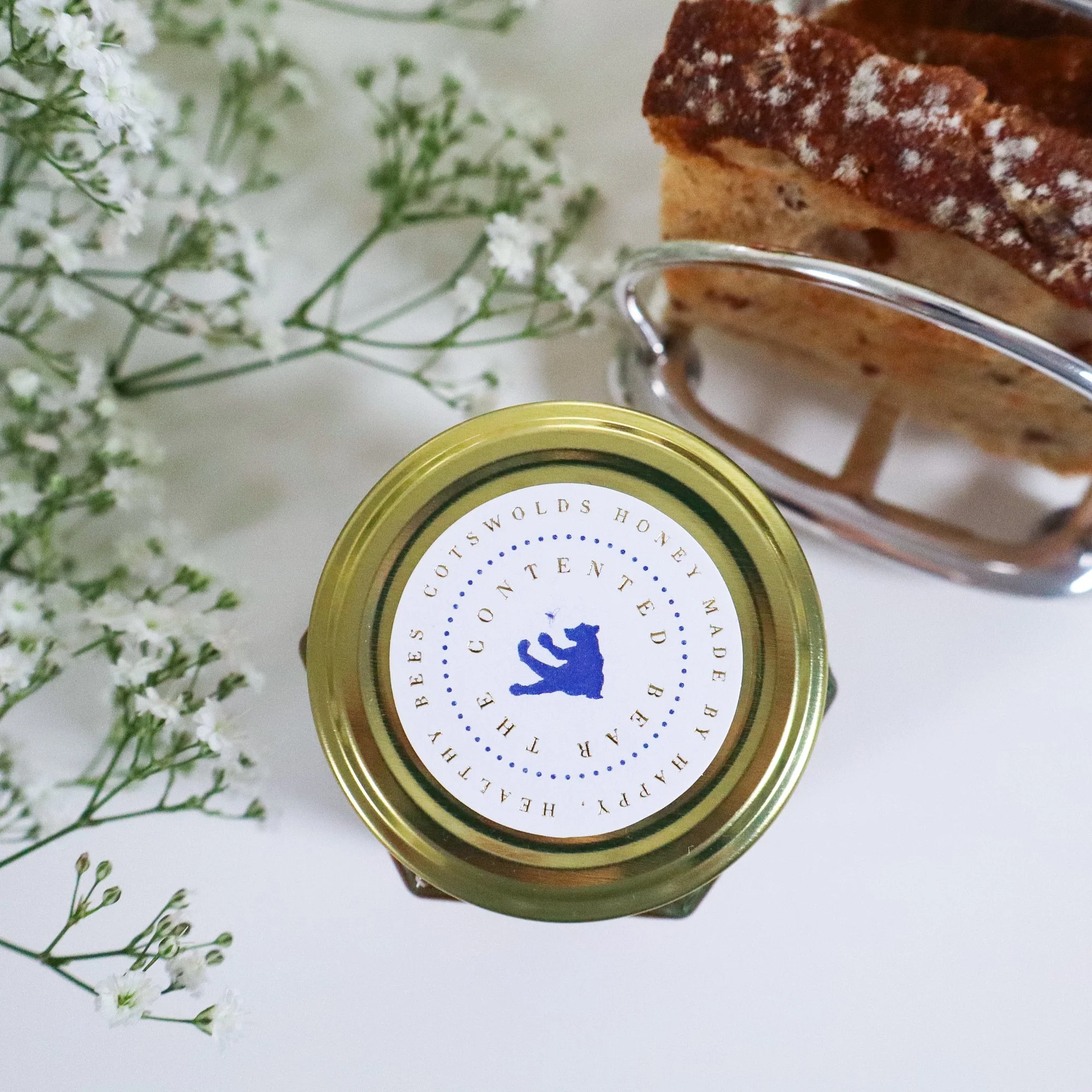 Beautiful over head view of a jar of luxury, sustainable honey with toast in a silver toast rack and white flowers.