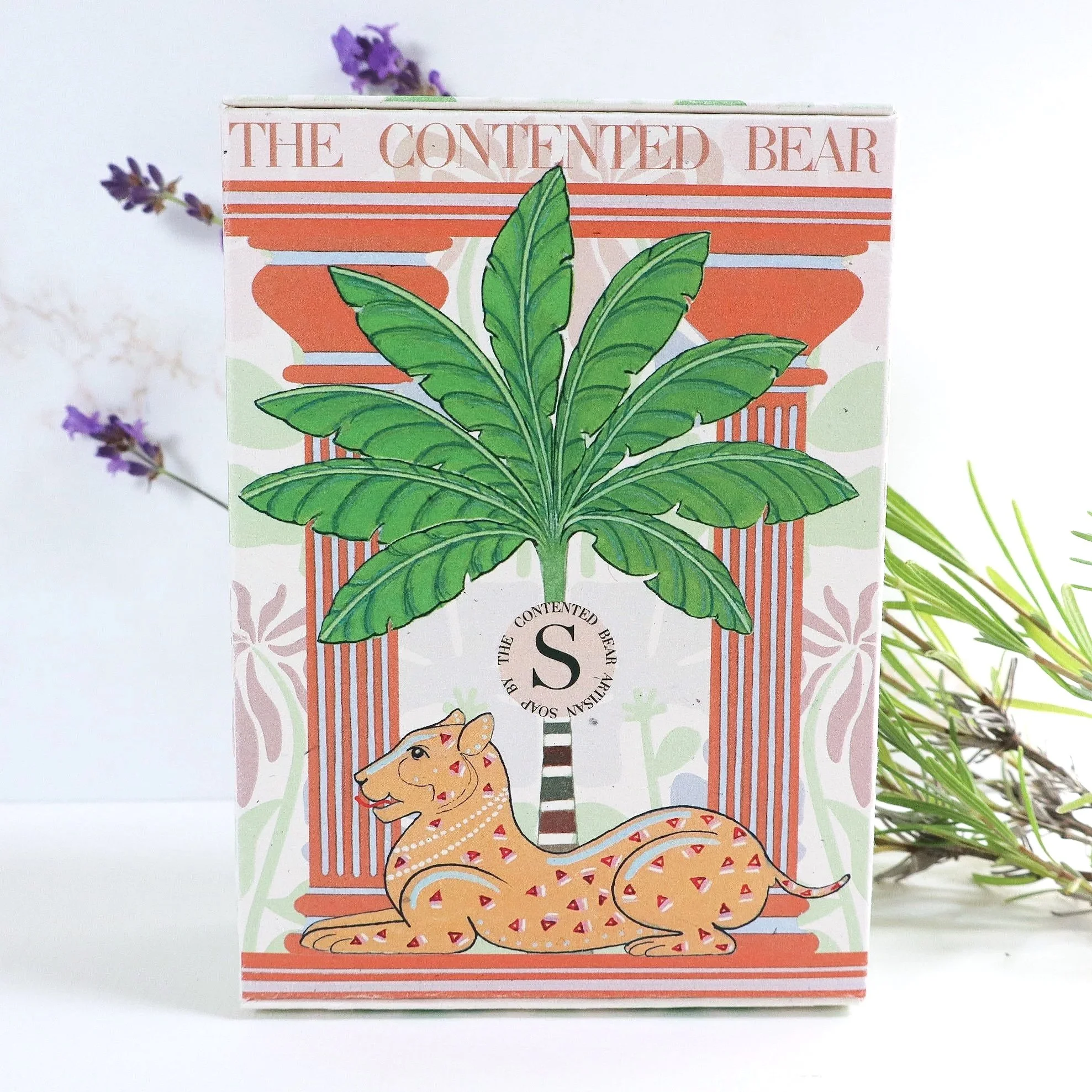 A photo of an eco luxury soap box featuring a hand painted palm tree and lioness in a temple garden. Fresh lavender stands up in the background.