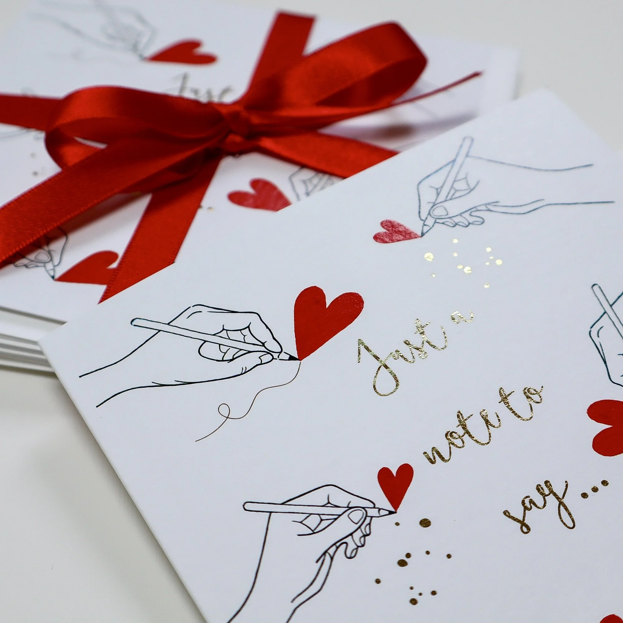 A close up of the red and white Just a note to say cards, featuring hands drawing hearts and gold foil doodles and ink.