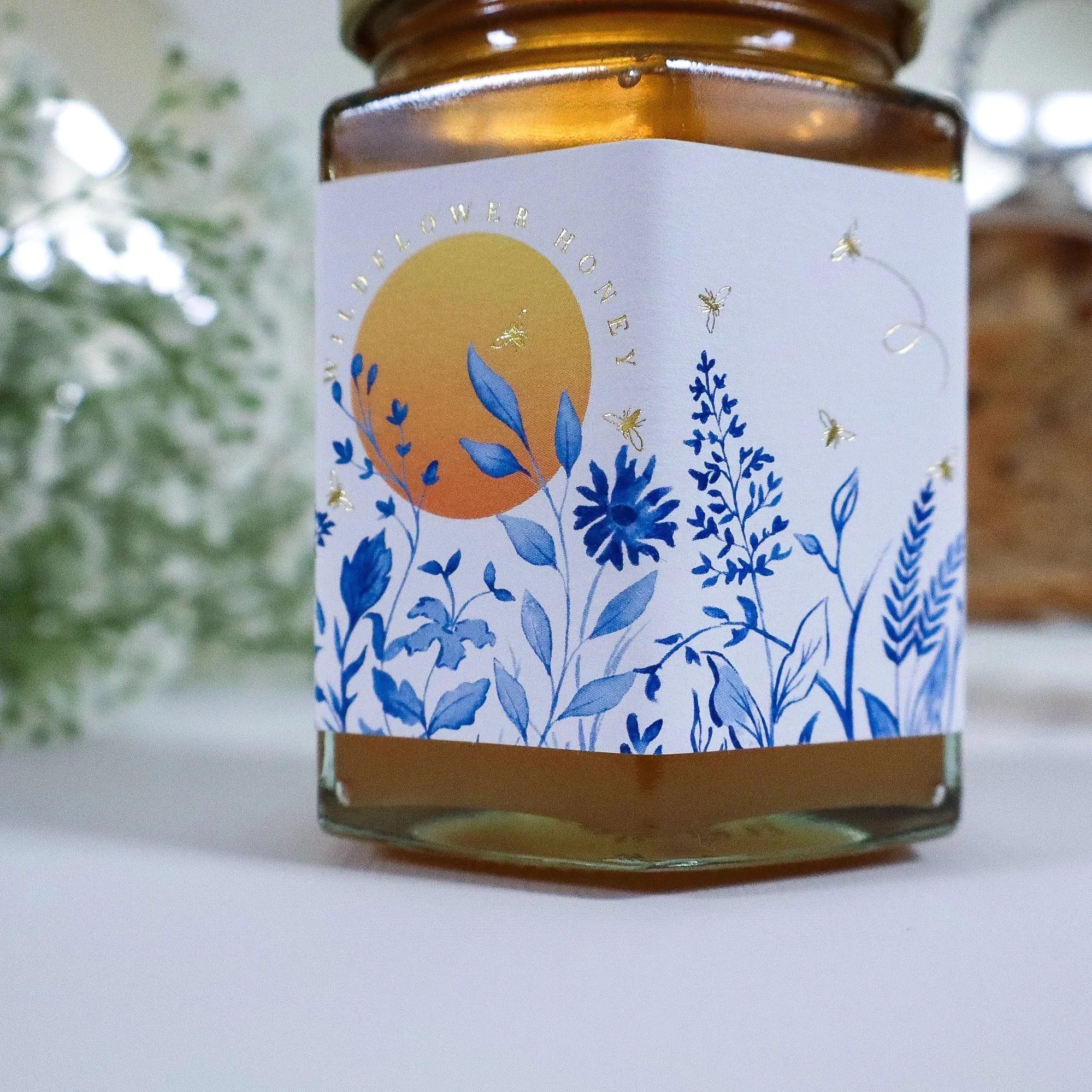 Side view of a jar of luxury, ethical honey made through kinder beekeeping. Luxury blue watercolour label of a meadow and sun with gold foil bees.