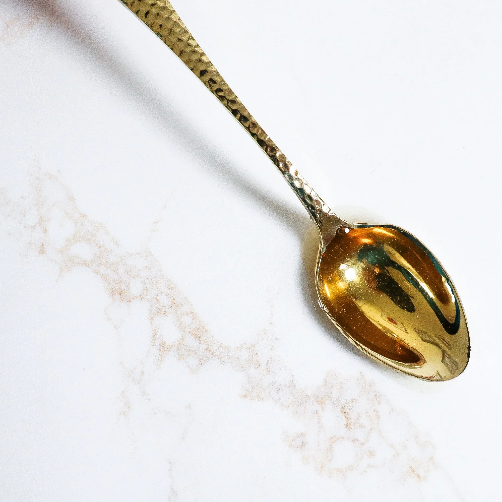 An aerial view of a golden teaspoon brimming with beautiful ethical honey, laying flat on a white marble kitchen top.