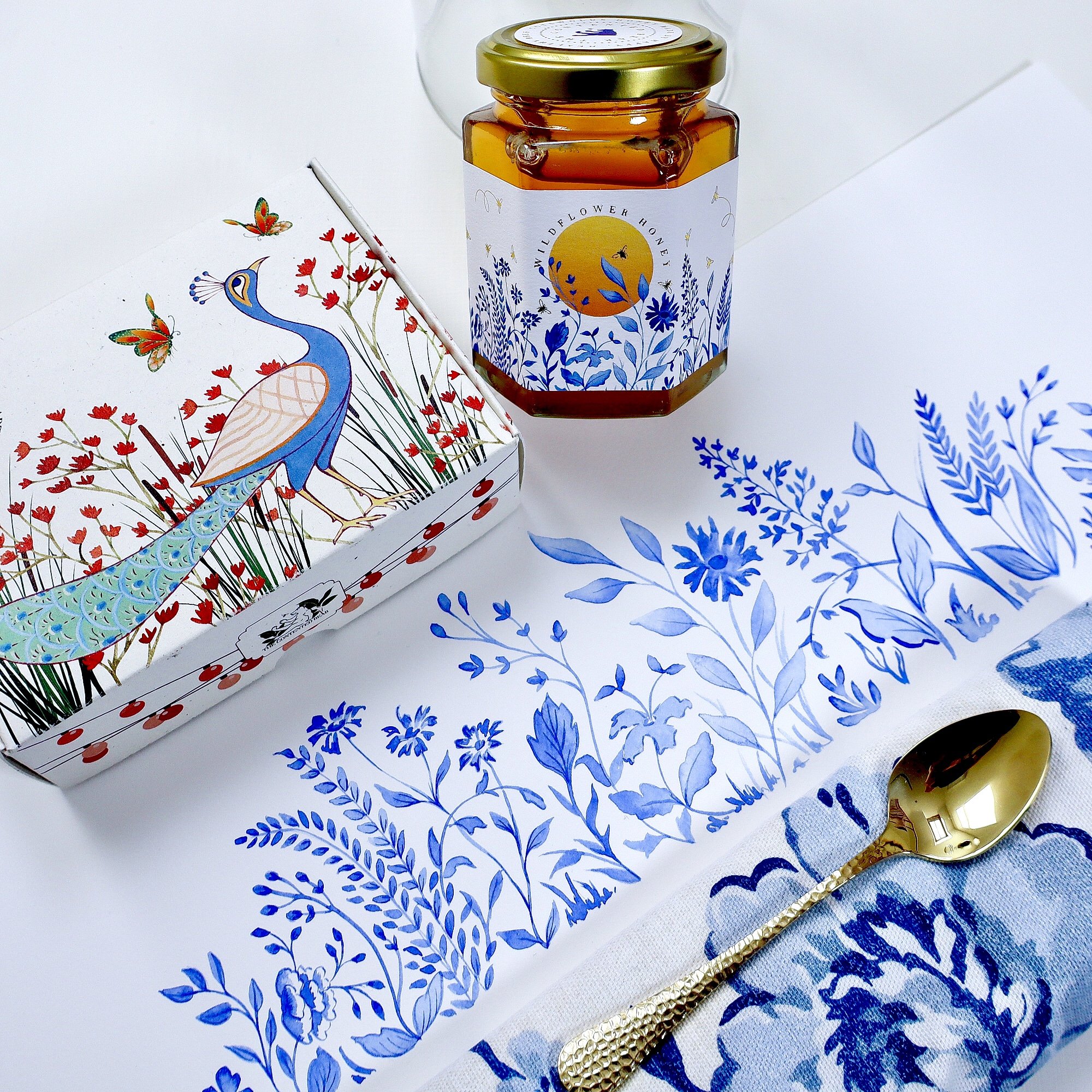 An aerial view of a watercolour of a wildflower meadow, linen napkin, peacock artisan soap box and a jar of ethical, luxury honey featuring the watercolour meadow. Everything is blue and white and gold. In the bottom right is a golden spoon sat atop