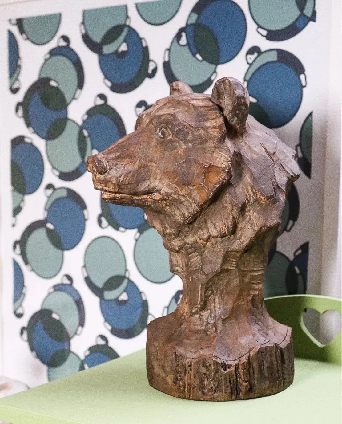A sculpted wooden  head of a bear with a kind smile looks out across a room. The room has a patterned background.