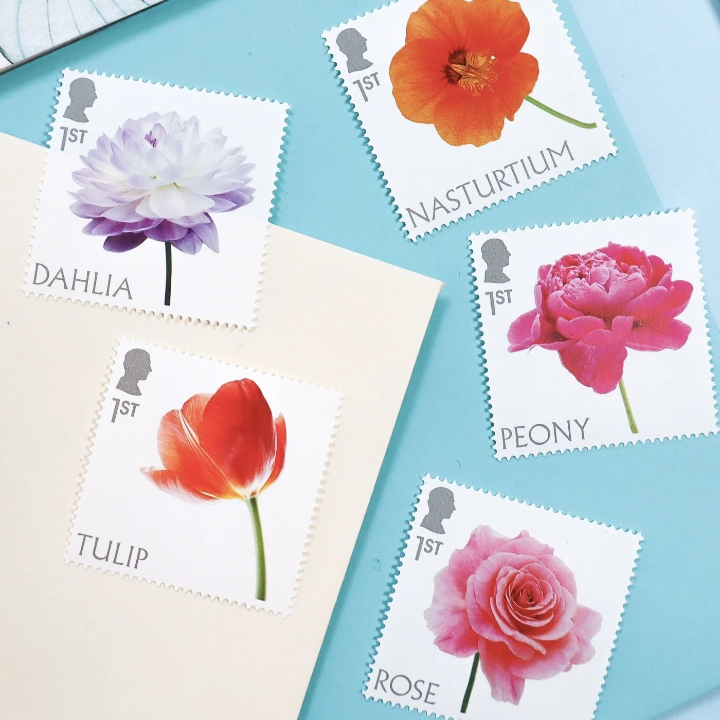 An aerial image of five stamps all featuring brightly coloured flowers on a background of a bright turquoise envelope and a cream envelope.