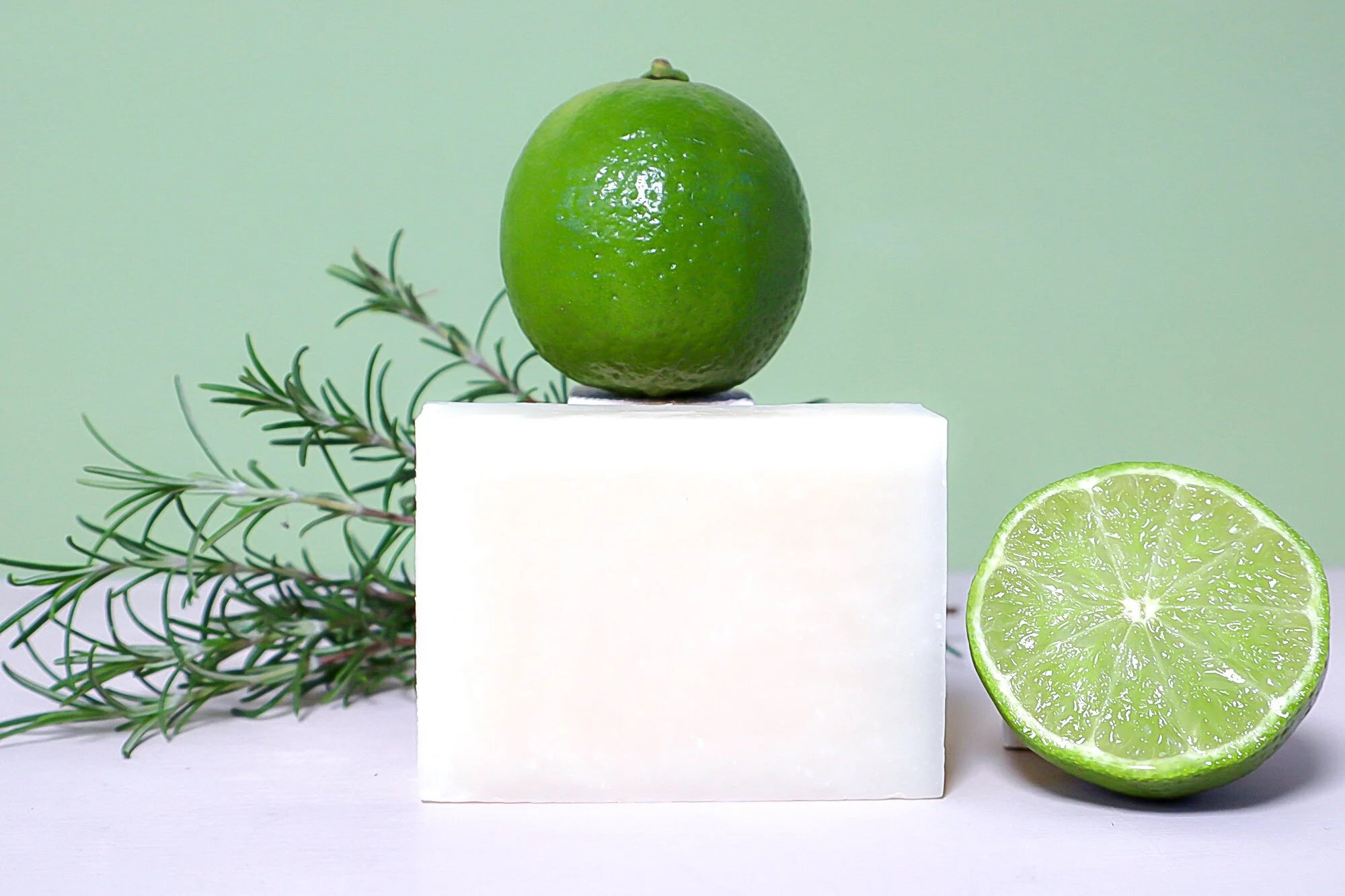 A beautiful photo of fresh, green limes with fresh bars of hand made soap and sprigs of fresh rosemary indicating the scent of the soap.
