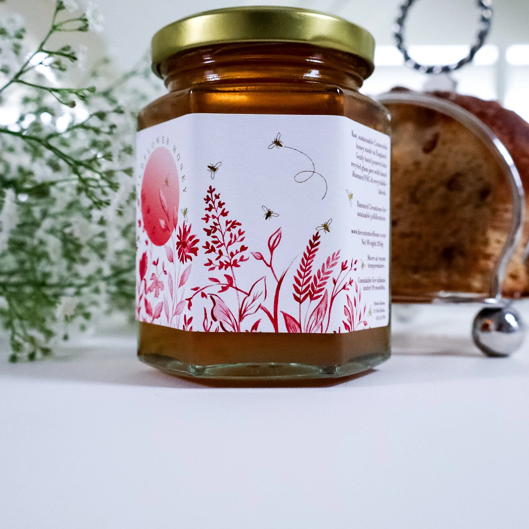 Side view of a beautiful, ethical, luxury jar of golden honey with premium hand illustrated label with gold foiling. To the left are delicate white flowers and to the right is a silver toast rack.