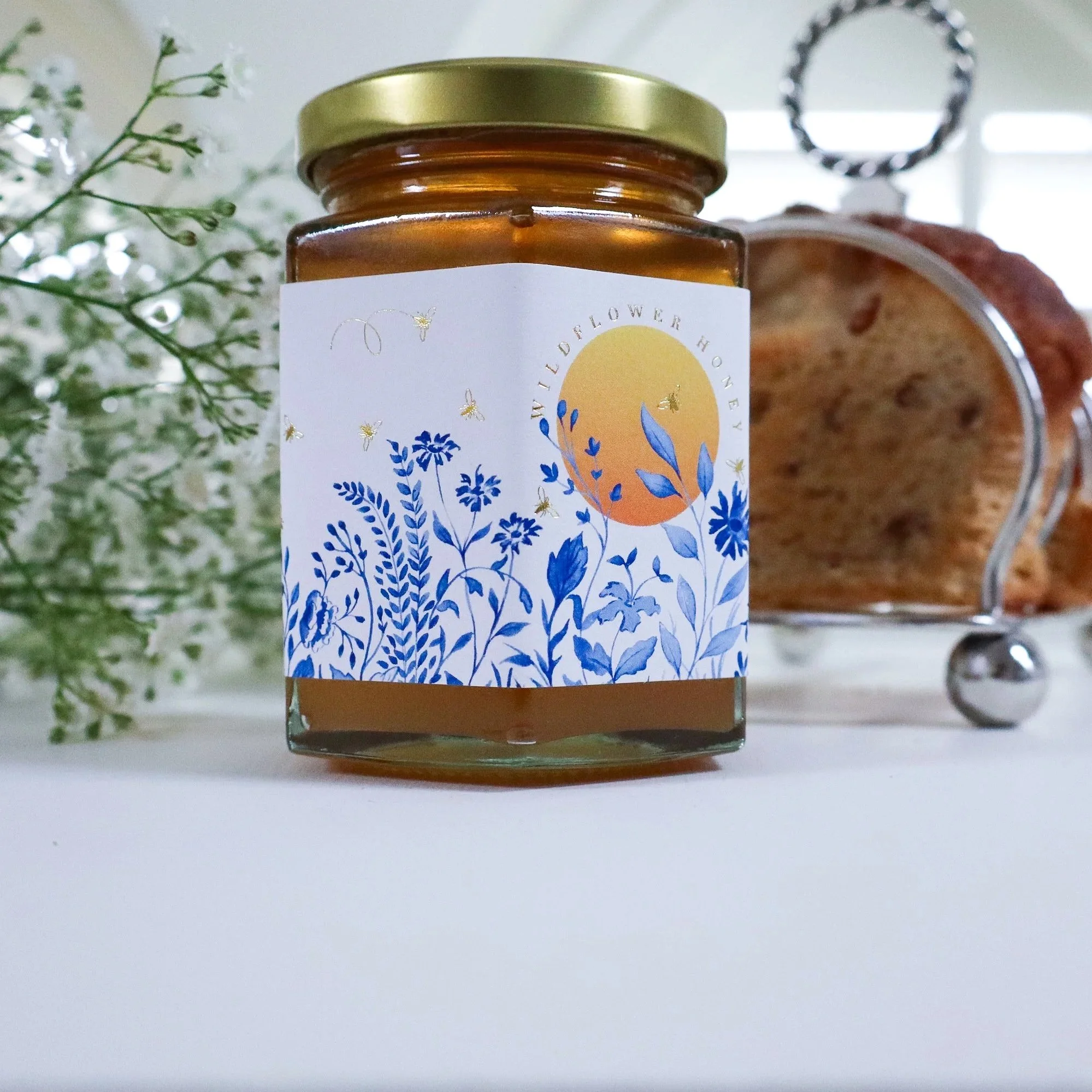 Side view of luxury, sustainable honey with a hand painted blue meadow scene and yellow sun adorning the label. Gold foil honeybees fly around the flowers.